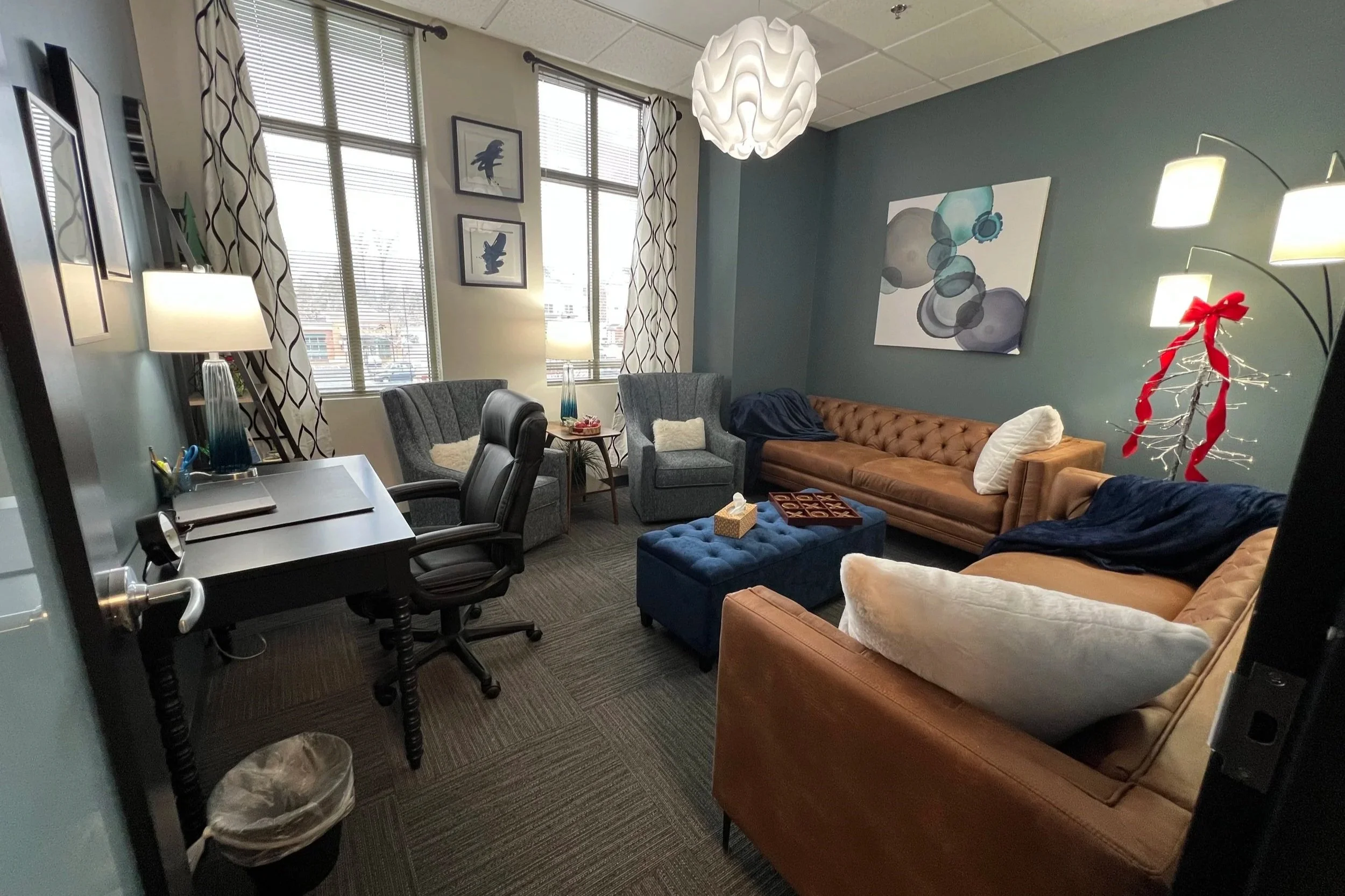 A cozy office waiting area with windows, curtains, framed artwork, a modern chandelier, lamps, armchairs, a brown leather tufted sofa, a blue ottoman with a tic-tac-toe game, a small side table with snacks, and a decorative towel holder with a red ribbon.