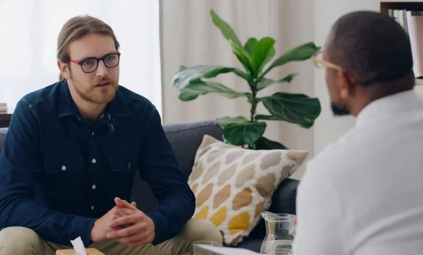 A man with glasses and light brown hair sitting on a sofa, engaged in conversation with another person wearing glasses. There is a large green plant and decorative pillows in the background.