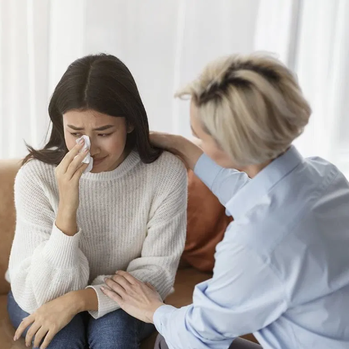 A woman with dark hair in a cream sweater crying and wiping her tears while a woman with short blonde hair in a light blue shirt comforts her, sitting on a couch in a bright room.