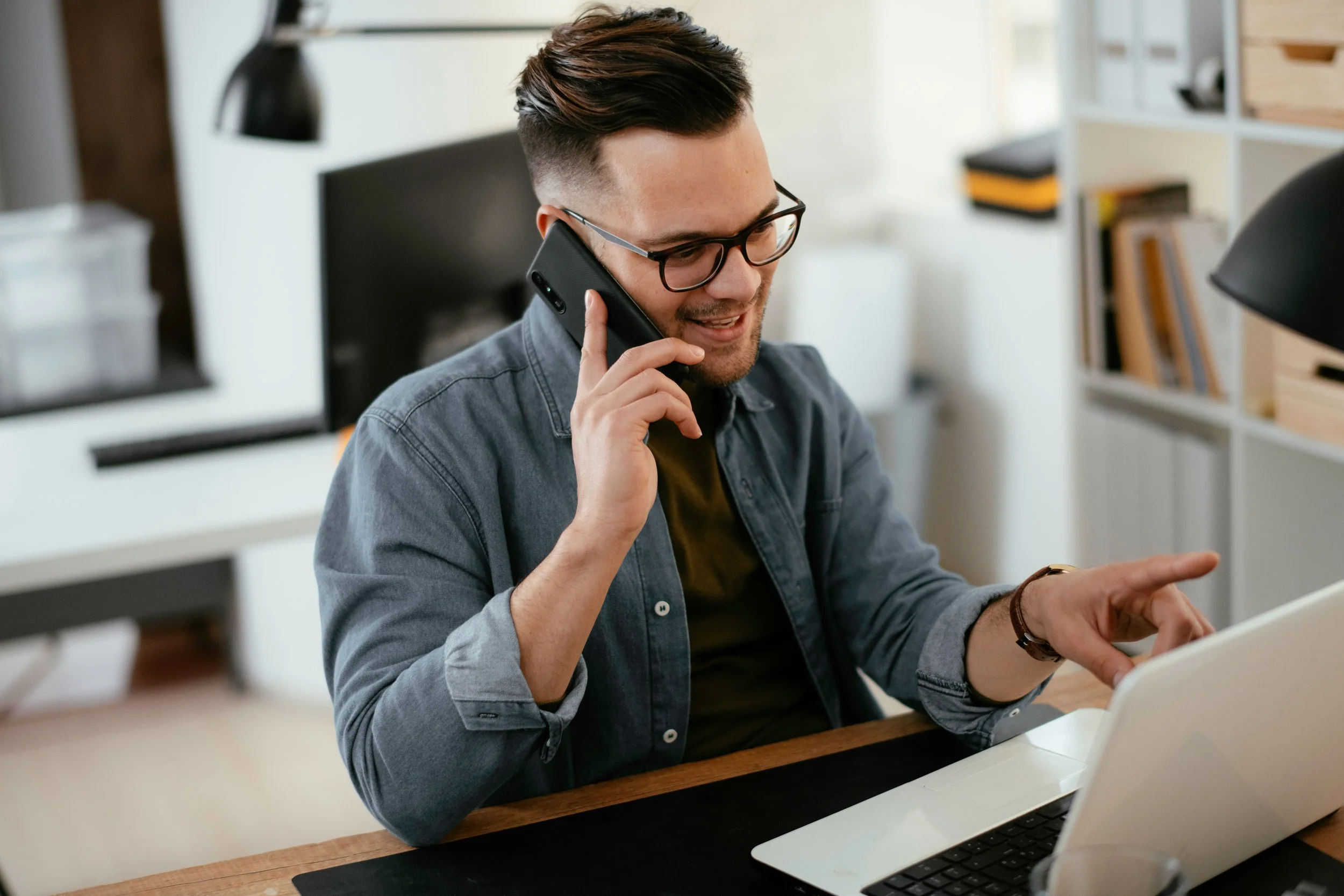 A young man with glasses, dark hair, and a beard, wearing a denim shirt, is sitting at a desk talking on a cellphone while using a laptop. The background features a modern office with shelves and a monitor.
