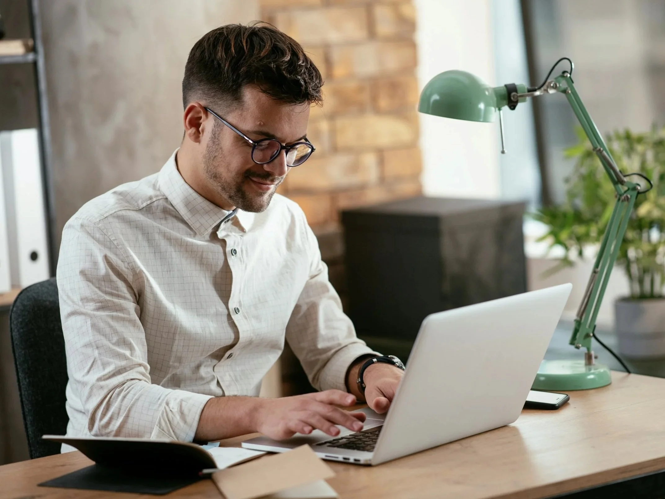 A young man with dark hair, glasses, and a beard, dressed in a white checkered shirt, working on a laptop at a desk. There is a green desk lamp and a smartphone on the desk. The background shows a brick wall and some plants.
