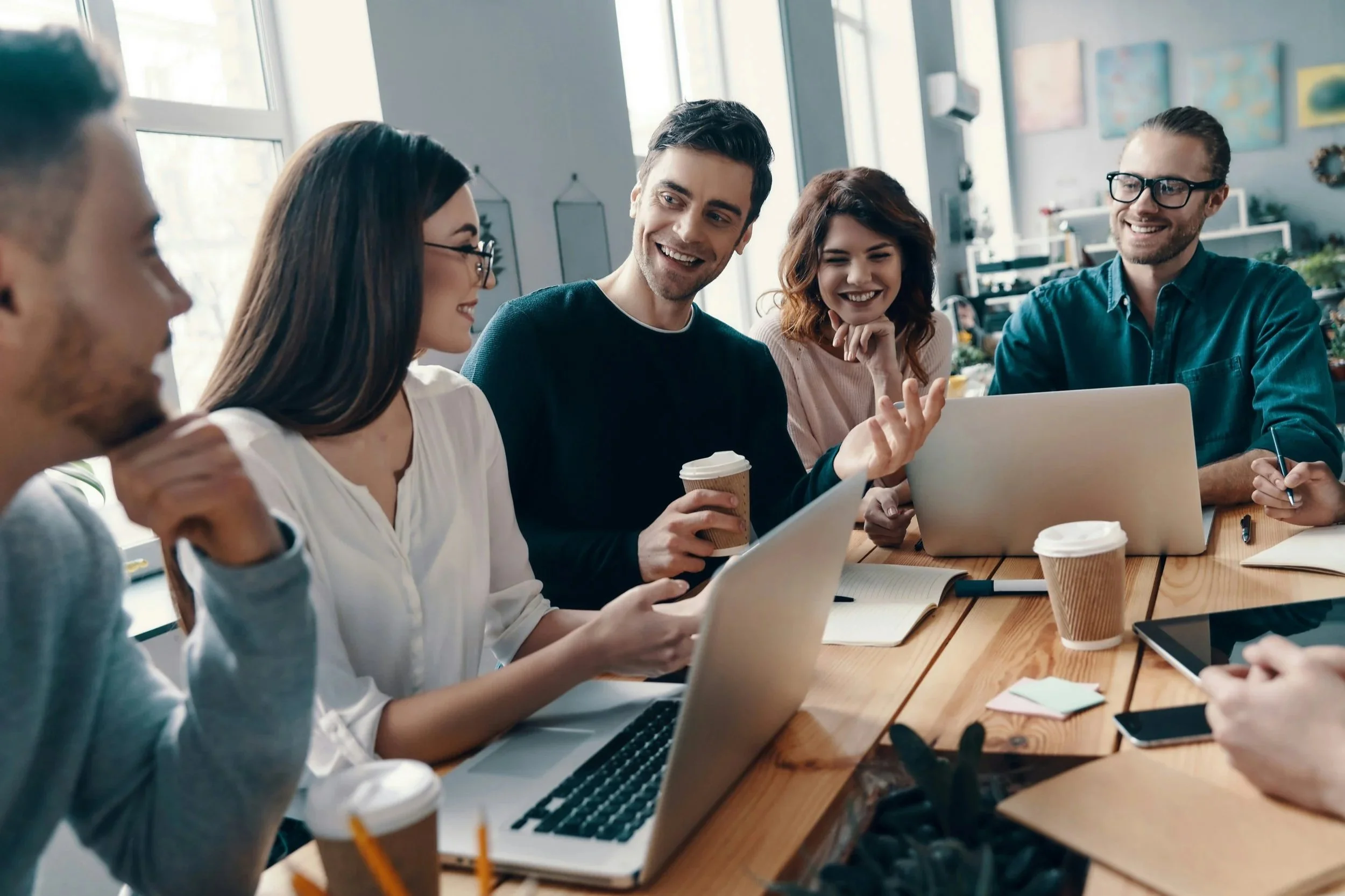A group of five young adults sitting around a wooden table engaged in a lively discussion, with laptops, notebooks, and coffee cups, in a bright office space with large windows and colorful artwork on the walls.