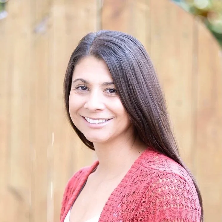 A woman with long dark hair smiling outdoors in front of a wooden fence, wearing a pink knitted sweater.