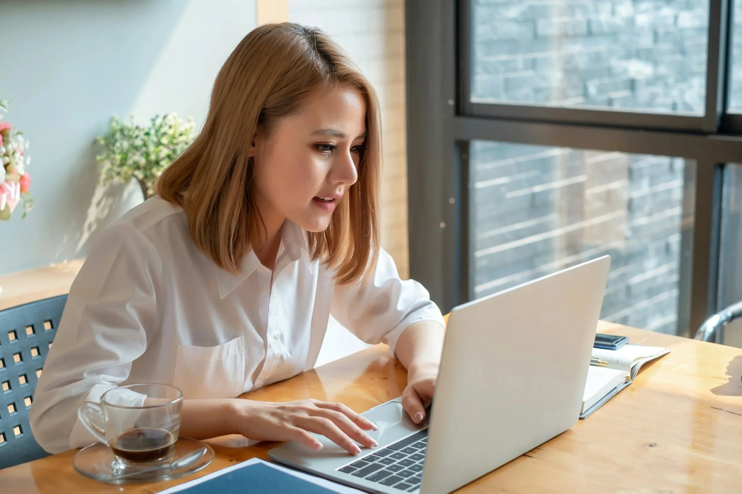 A woman with shoulder-length light brown hair working on a laptop at a wooden table, with a glass of dark soda and an open notebook nearby, in a bright room with large windows and a city view.