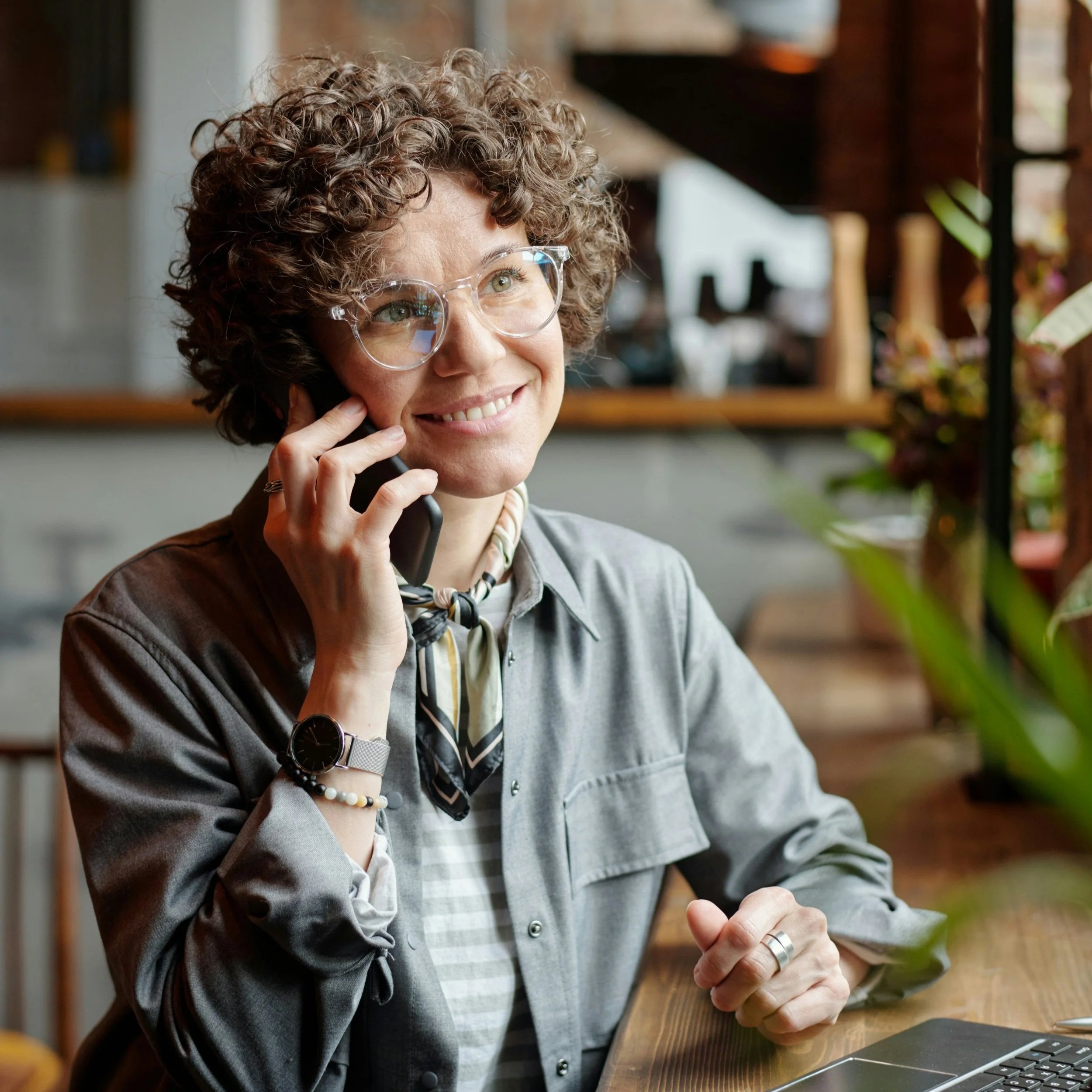 A woman with curly hair wearing glasses talking on a phone, smiling, while sitting at a wooden table with a laptop and plants in the background.