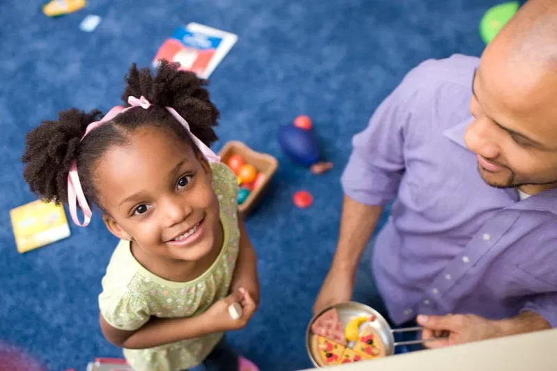 A young girl with puffy hair tied with pink ribbons, smiling and looking up at the camera, while a man in a purple shirt holds a game or toy with a pizza slice and other objects, and there are scattered toys and books on a blue carpeted floor.