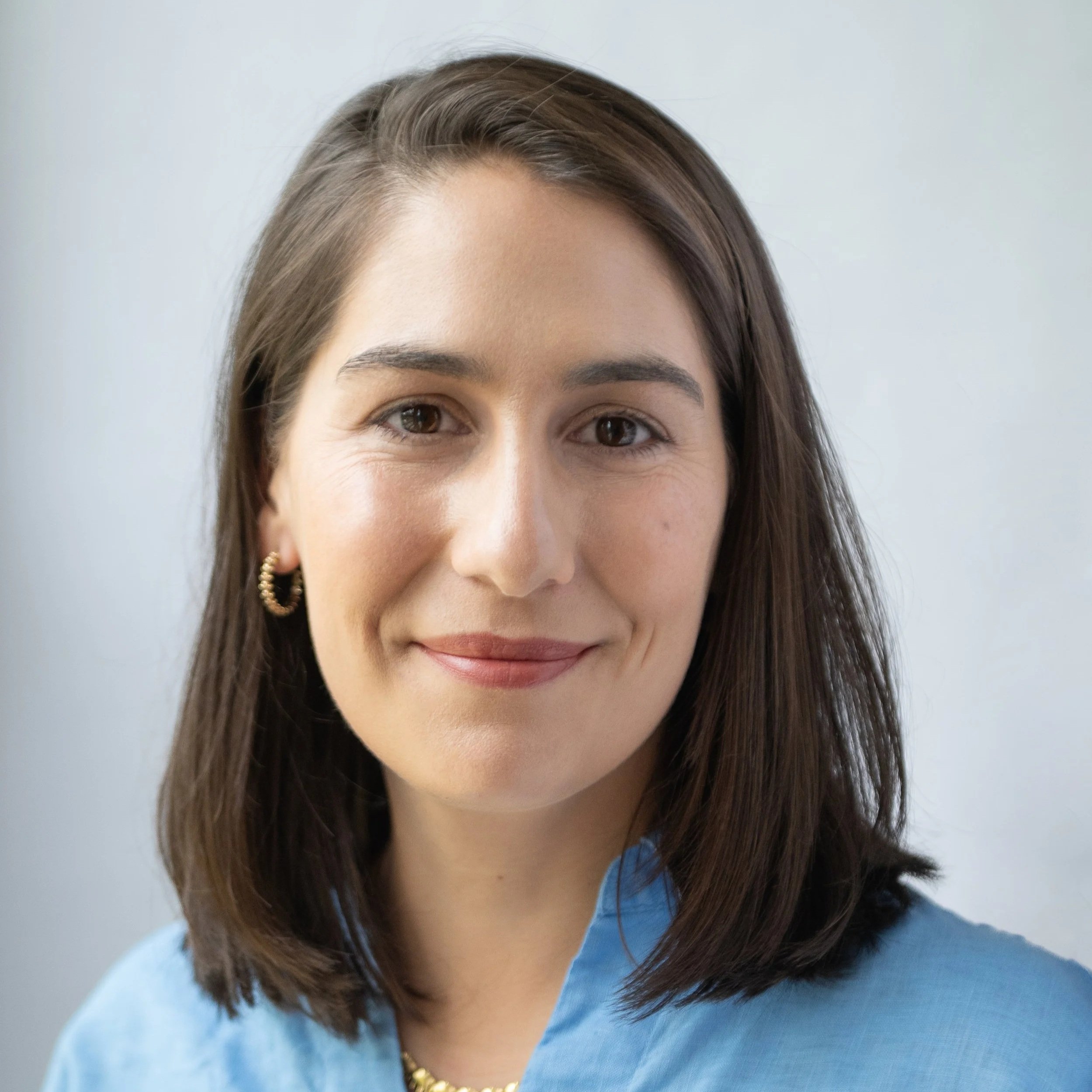 Close-up of a woman's face with smile, brown hair, and gold hoop earrings, wearing a light blue top, against a plain background.
