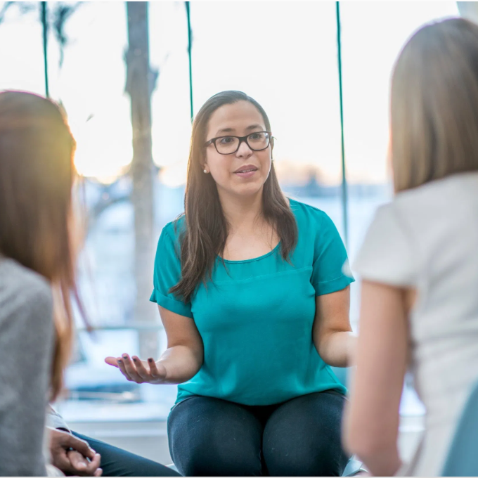 Woman in teal shirt and glasses speaking and gesturing with her hands while seated with two other women in a brightly lit room with large windows and a view of boats or water outside.