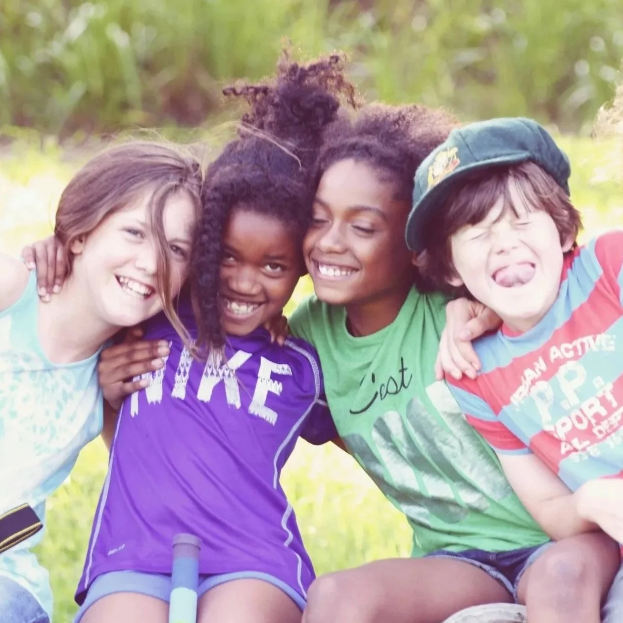 Four kids smiling and hugging outdoors in a grassy area.