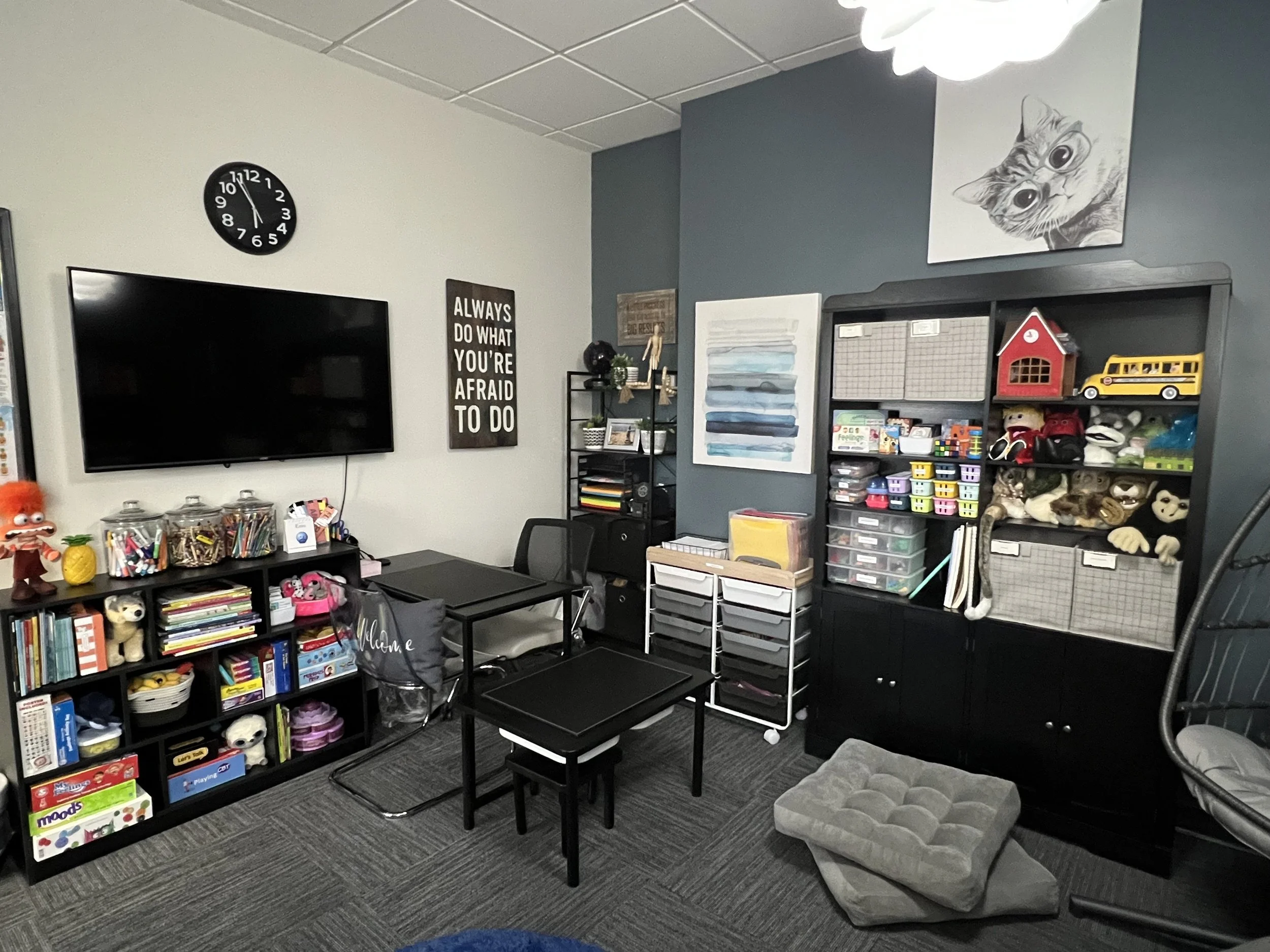 A colorful classroom corner filled with toys, books, and art supplies, including plush animals and storage bins, with a clock on the wall and a black shelf holding various items.