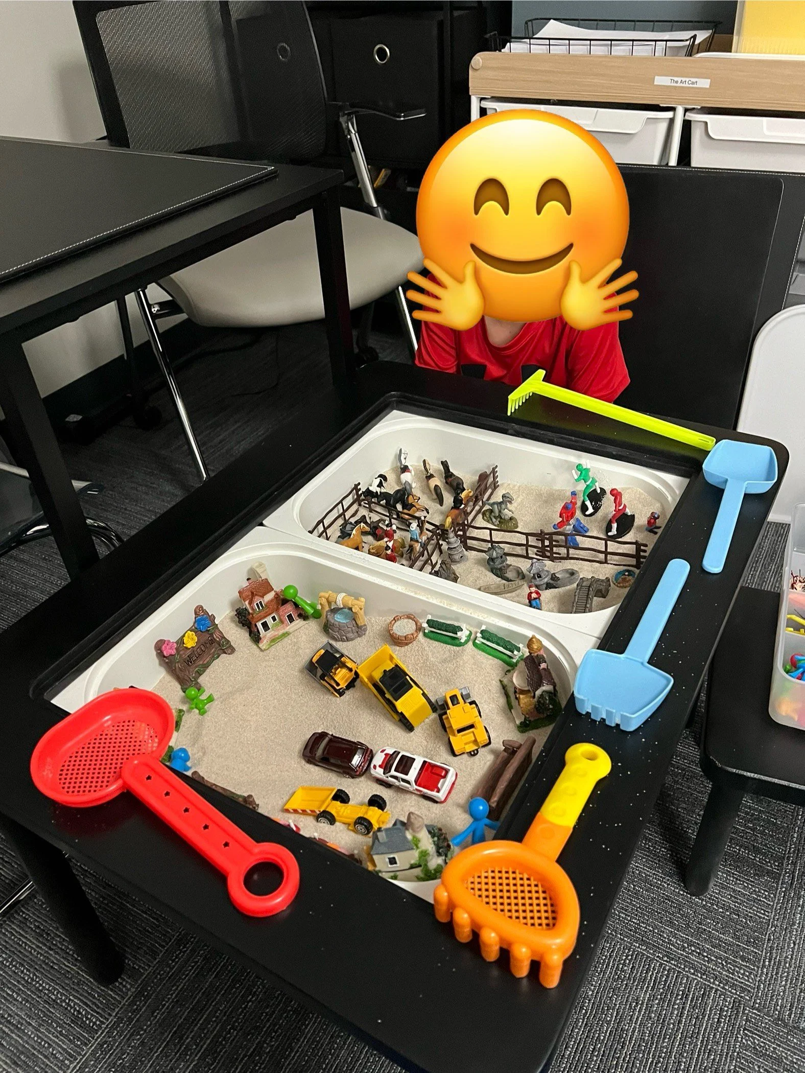 A black table with a sandbox filled with toy cars, animals, and miniature structures; various colorful toy scoops and tools are visible around the sandbox.