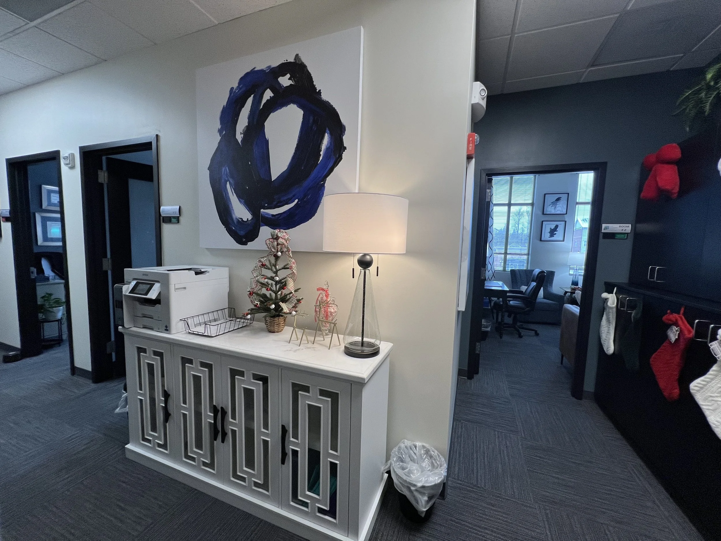 An office reception or waiting area with a white decorative cabinet in the center, topped with a lamp, a small Christmas tree, and decorative items. Behind the cabinet, there is a modern, abstract painting with black and blue colors. To the left are two doorway openings with dark frames. On the right, a room with window and framed bird pictures on the wall is partially visible. The floor is carpeted with dark gray tiles, and there are various office equipment and holiday decorations around.