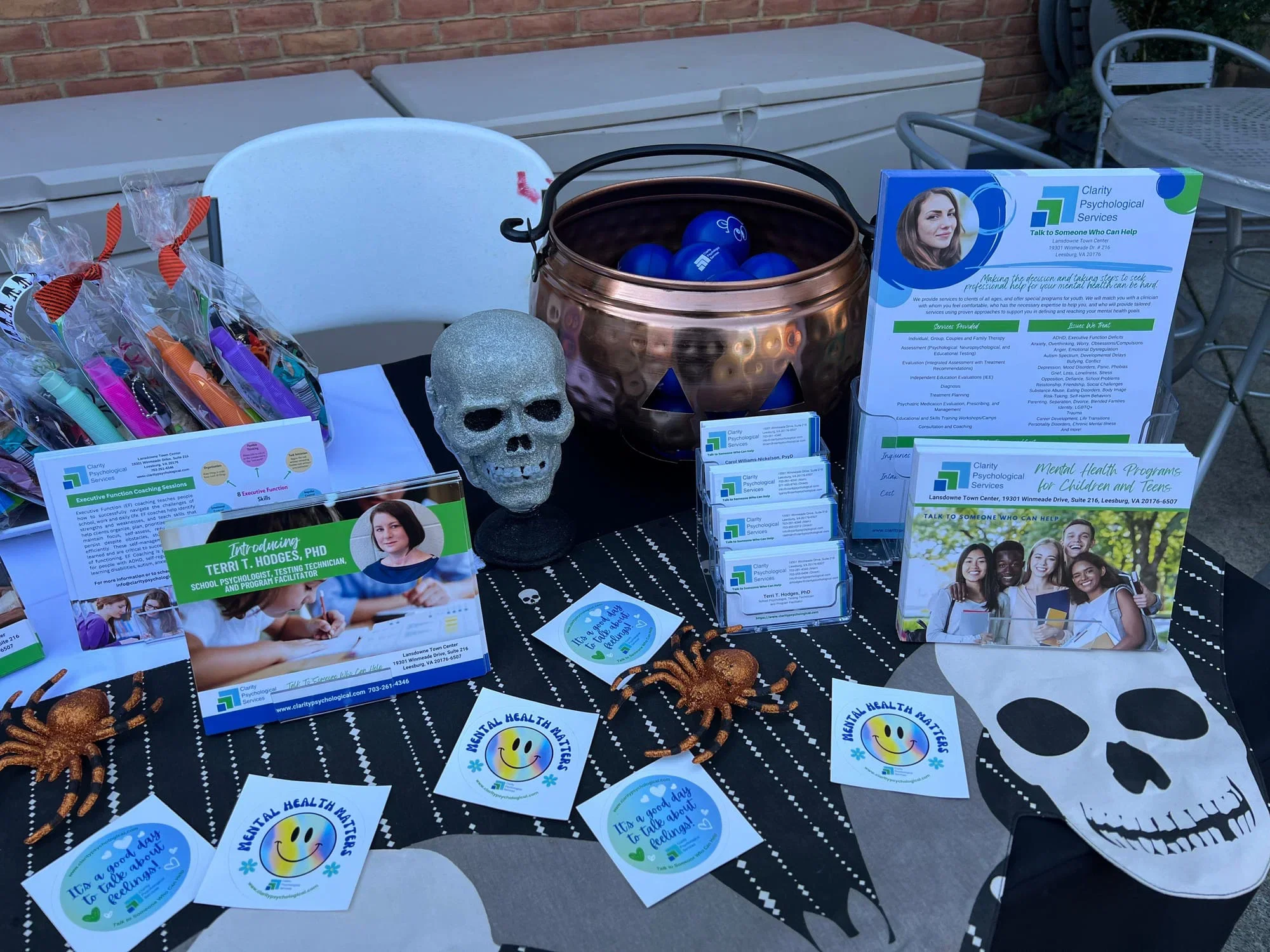 An outdoor table with informational flyers, business cards, and small decorative items related to mental health services, including a skull and fake spiders as decorations.
