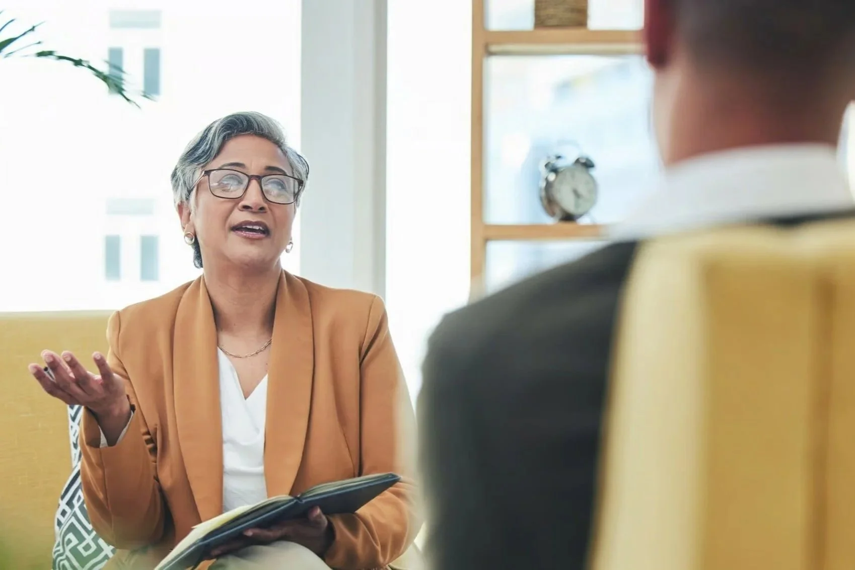 An older woman with short gray hair, glasses, and wearing a tan blazer is speaking during a conversation or interview inside a bright room, with a person in a dark suit listening to her.