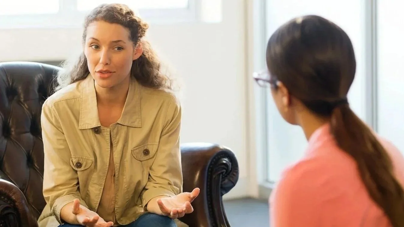 Two women having a conversation in an office setting. One woman, with curly hair and wearing a beige jacket, is gesturing with her hands. The other woman, with dark hair tied back and wearing glasses and a pink top, is listening.