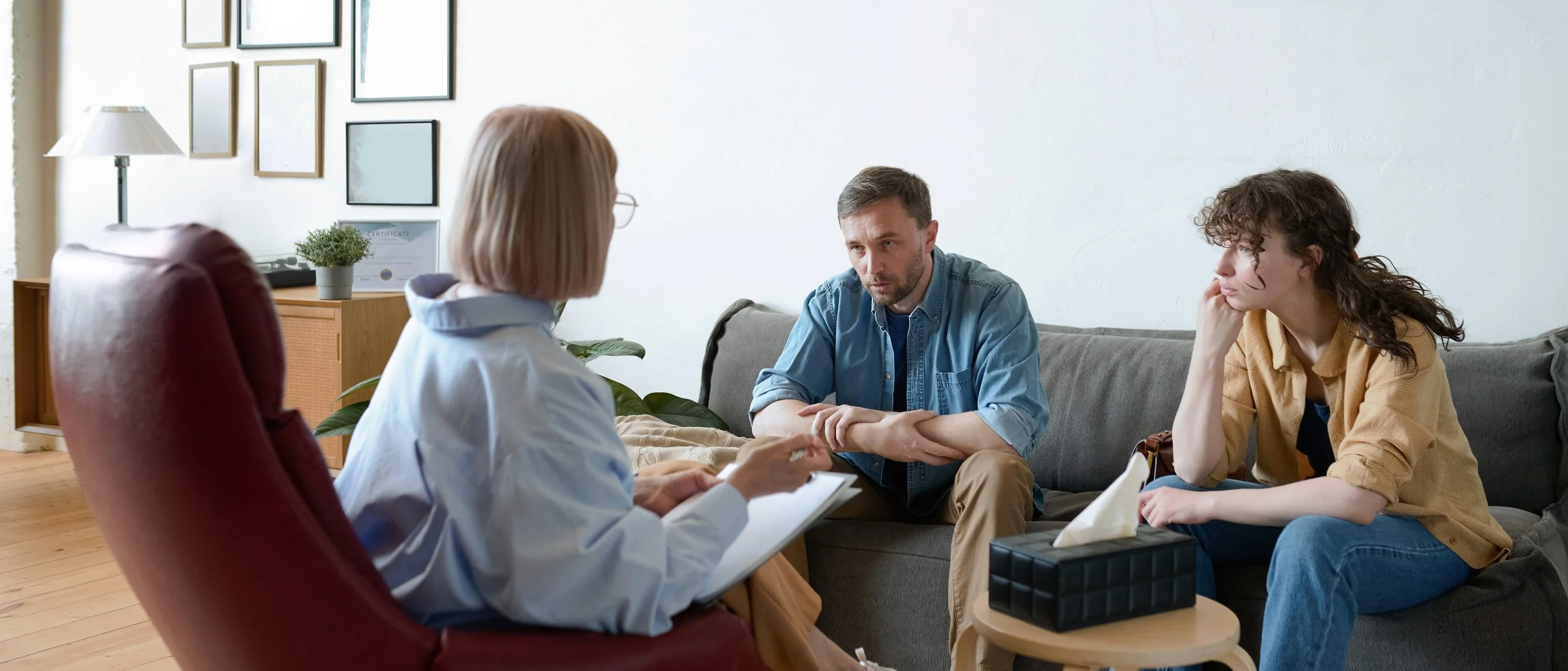 A therapist or counselor sitting on a red chair with a notepad, talking to a man and a woman seated on a gray sofa in a therapy session or counseling session.