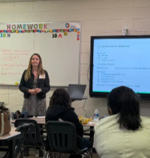 A teacher standing in front of a classroom with students, presenting a lesson on a large screen. The classroom has a whiteboard with 'HOMEWORK' written on it and colorful decorations.