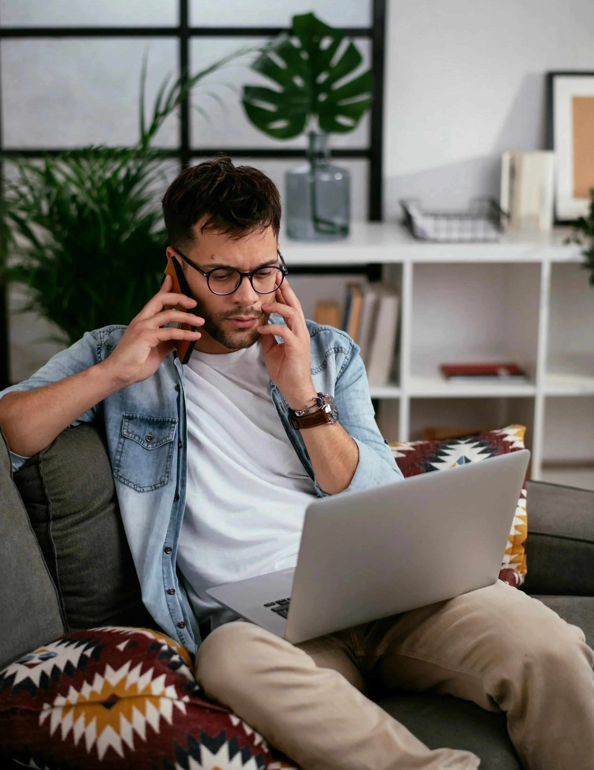A man with glasses and a beard, wearing a denim shirt over a white T-shirt, sitting on a couch, talking on the phone with a laptop on his lap, in a modern living room with houseplants and bookshelves.