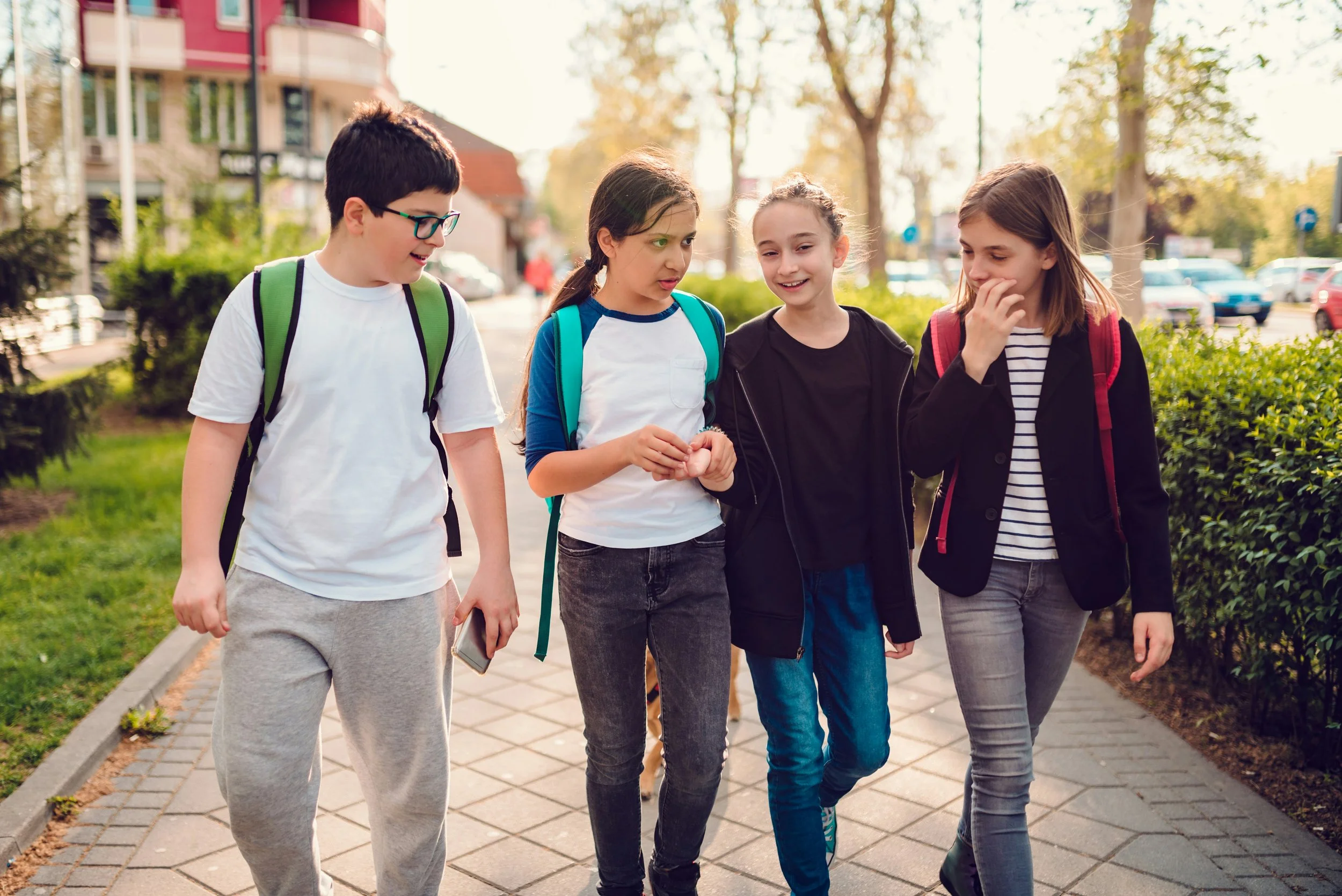 Four kids walking together outdoors on a sidewalk, talking and smiling, with trees and parked cars in the background.