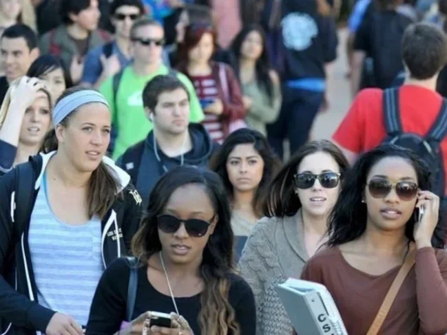 A crowd of young adults walking, some wearing sunglasses, with a diverse group in an outdoor setting.
