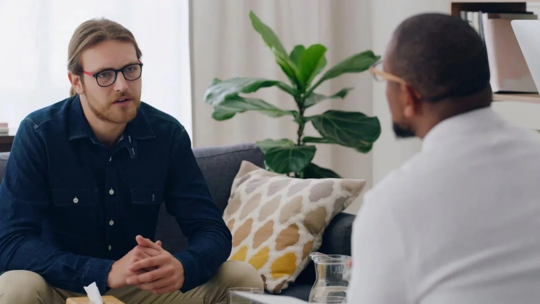 Two men seated on a sofa having a serious conversation. One man has brown hair, glasses, and a beard, wearing a dark blue shirt. The other man has dark hair, glasses, and a goatee, wearing a white shirt. A large green houseplant and a cushion with a yellow and white pattern are visible in the background.