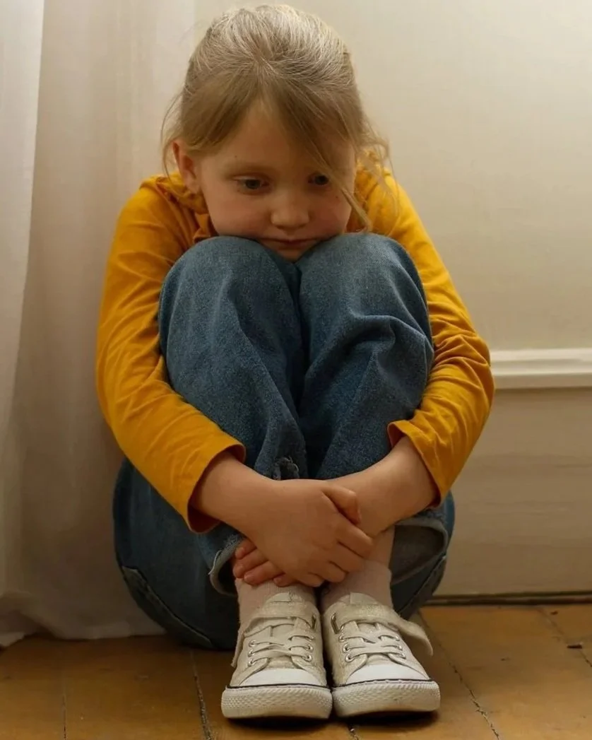 A young girl sitting on the floor with her knees pulled to her chest, hugging her legs, wearing a yellow long-sleeve shirt, jeans, and white sneakers, with a neutral expression.