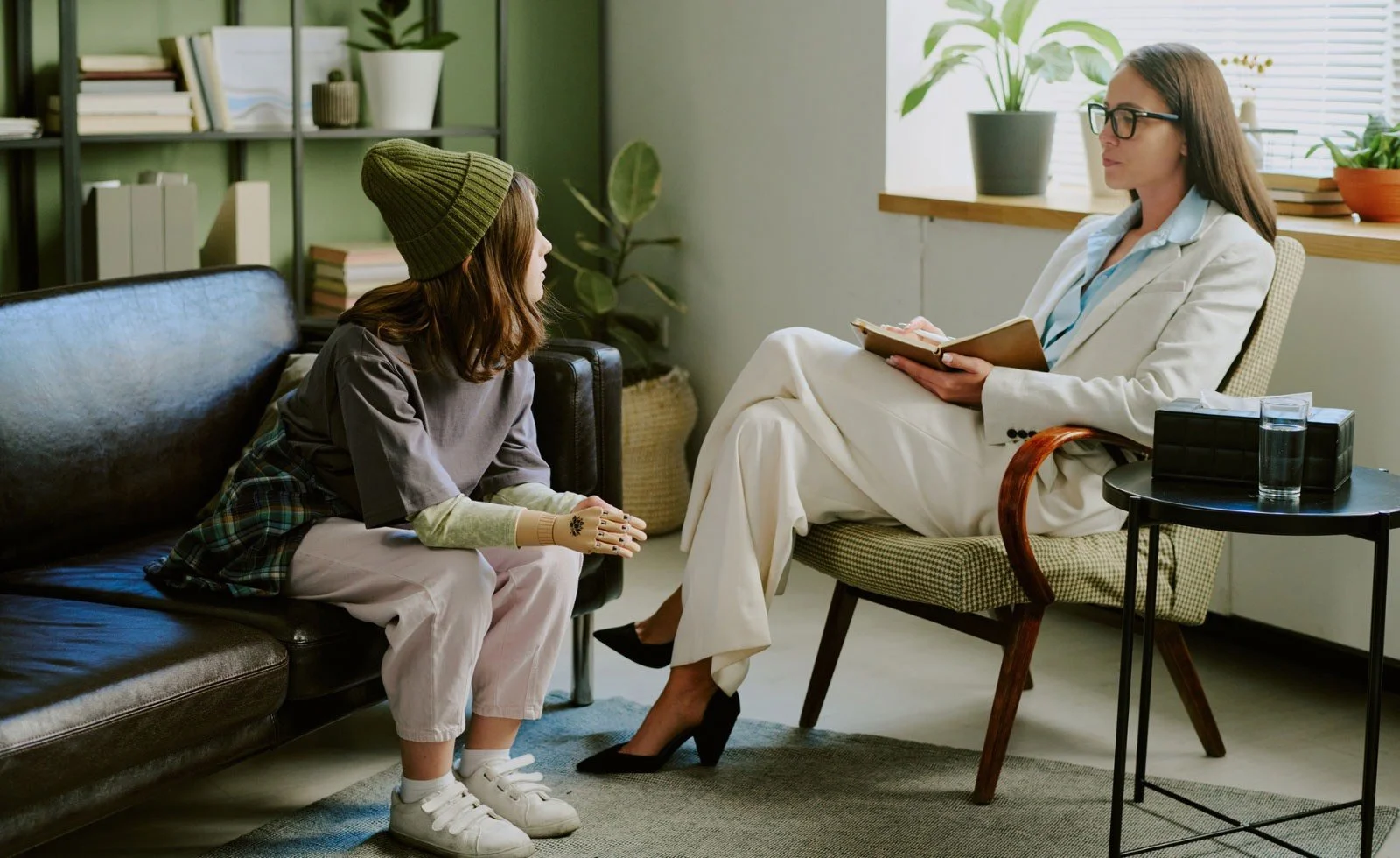 A young woman sitting on a black leather sofa during a therapy session, speaking with a female therapist who is sitting on a chair, holding a notepad, in a well-lit room with plants and books.