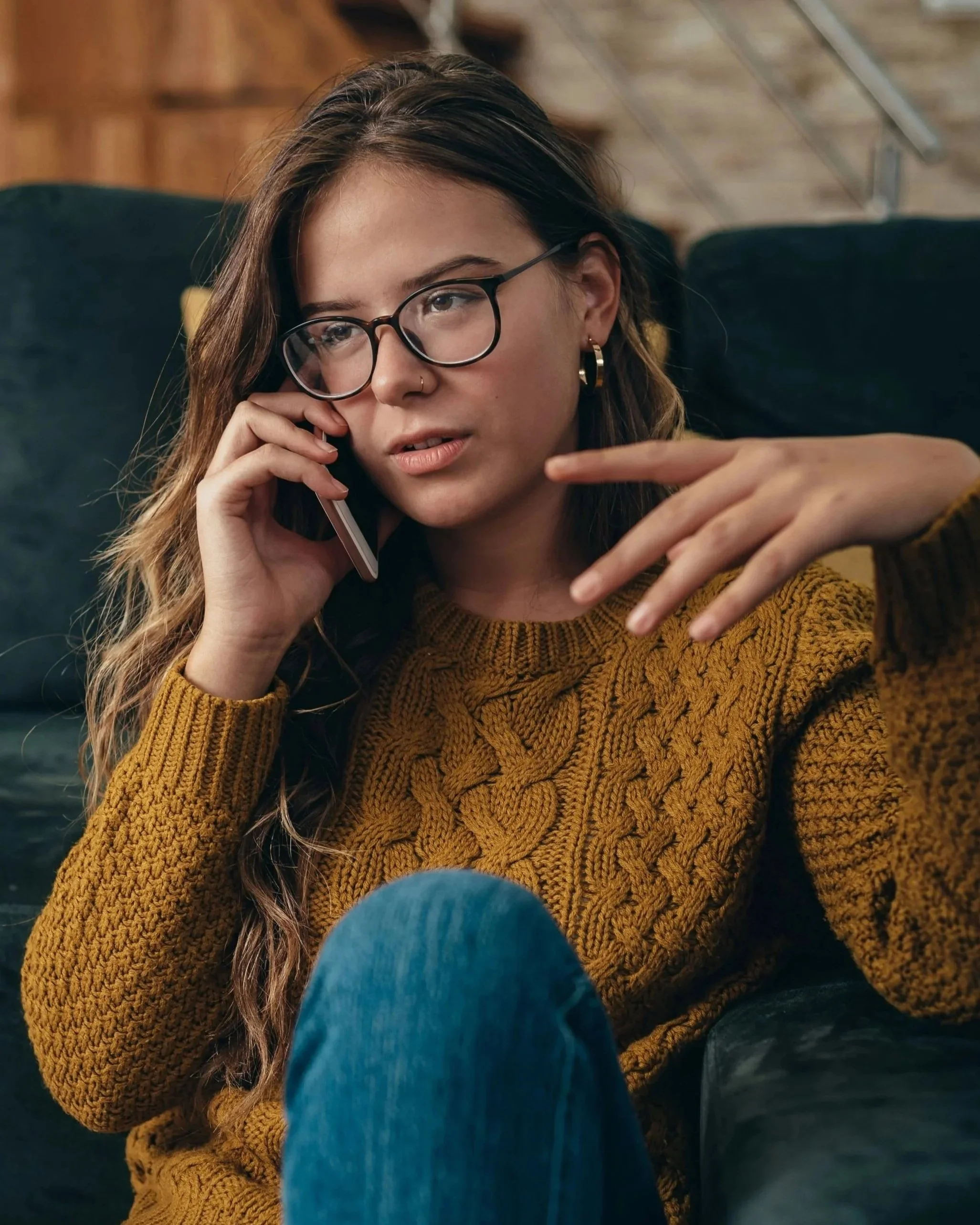 Close-up of a young woman with long brown hair, wearing glasses, a nose ring, gold hoop earrings, and a mustard-colored knit sweater, sitting on a dark couch, looking thoughtful while talking on her phone. In the background are wooden shelves and a metallic ladder.