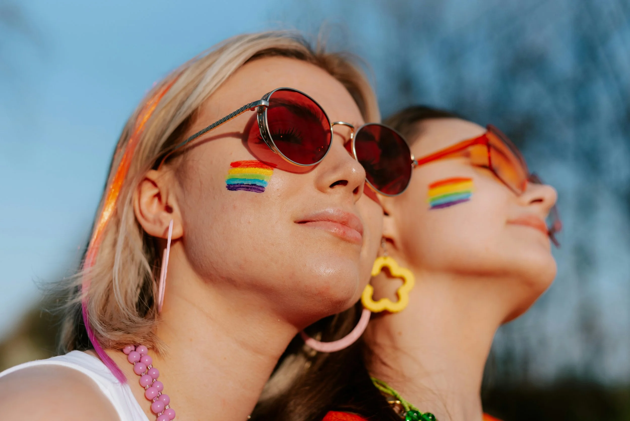 Two women with rainbow flag face paint, wearing sunglasses, colorful accessories, and smiling outdoors.