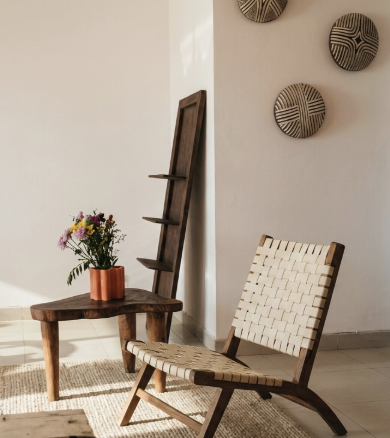 A cozy interior corner with a wooden chair featuring a woven seat and backrest, a small wooden side table with a potted flower bouquet, a tall wooden leaning shelf with two small shelves, and three decorative wall hangings with tribal designs.