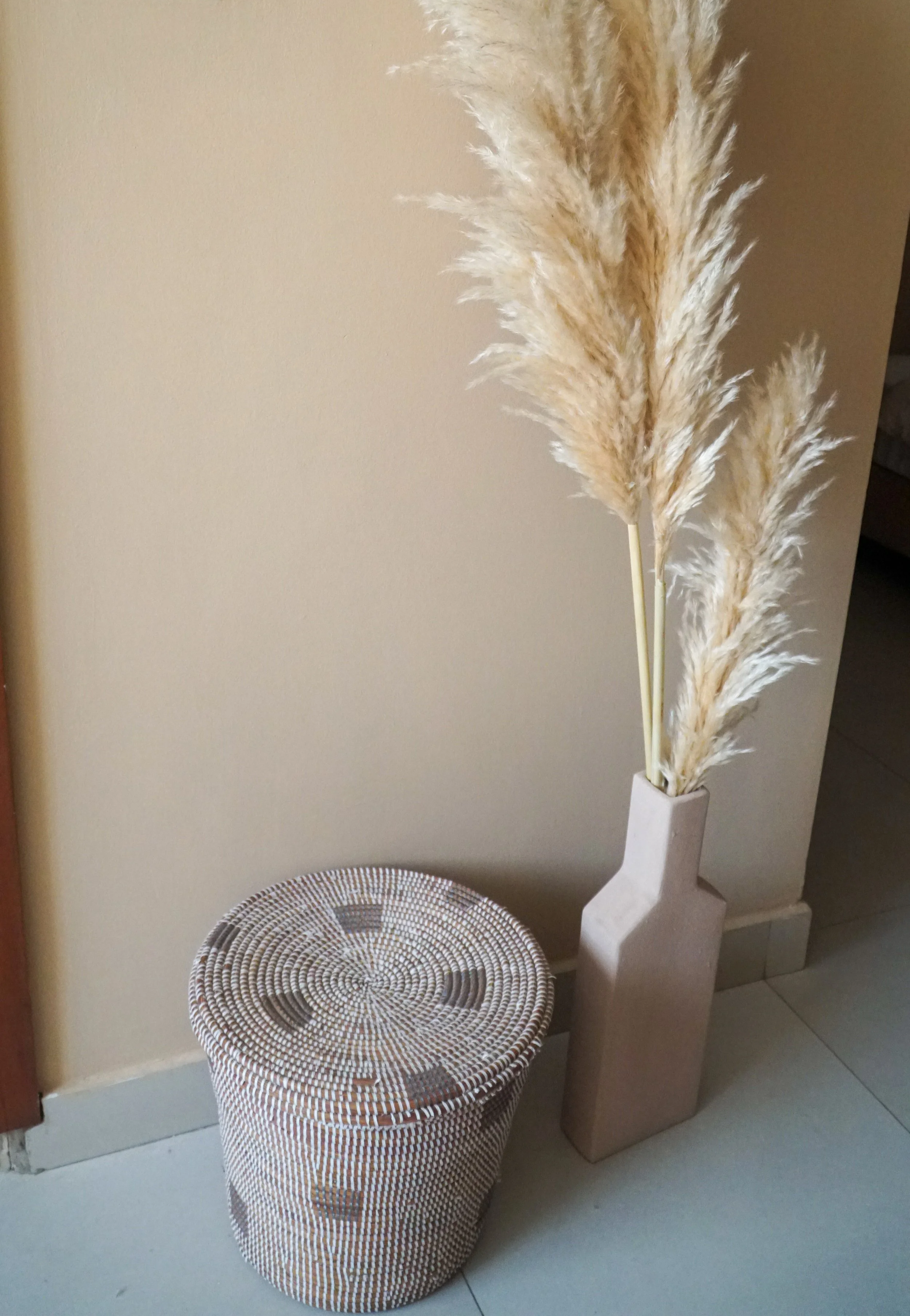 A beige vase with dried pampas grass next to a round woven basket with geometric patterns, placed against a neutral-colored wall.