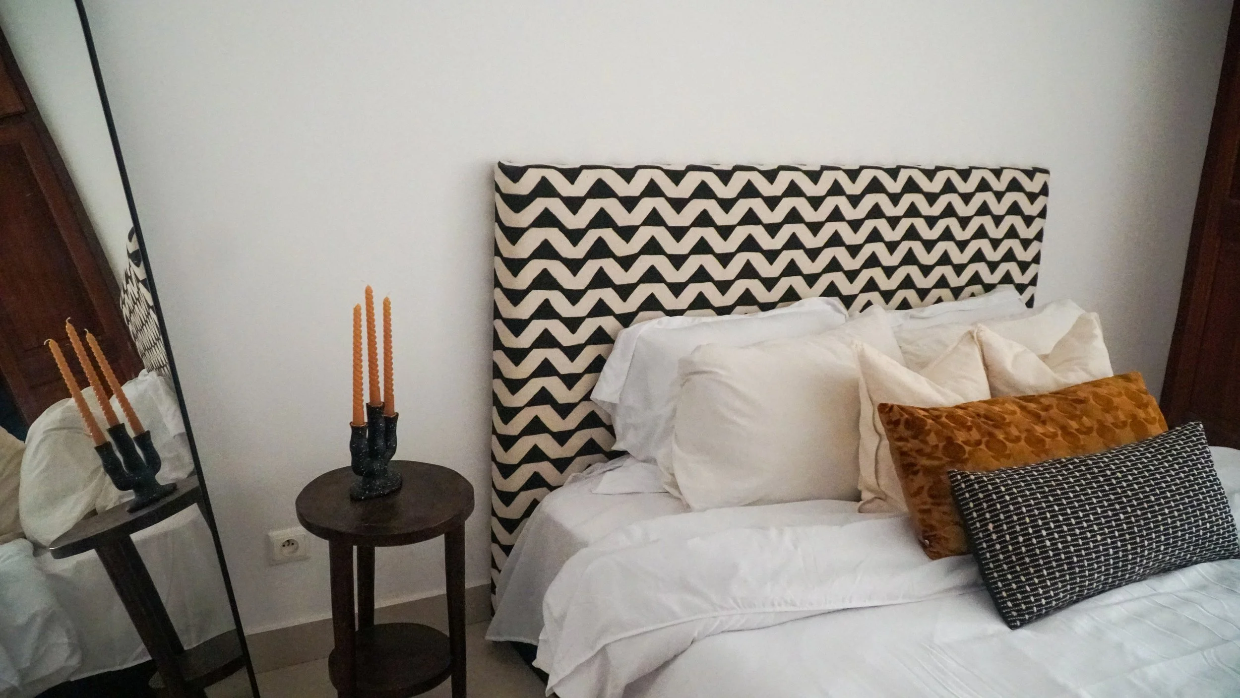 A bedroom with a bed featuring a black and white chevron-patterned headboard, multiple white pillows, and decorative pillows in brown and black patterns. A round wooden side table with a black candle holder and orange candles is next to the bed. A la
