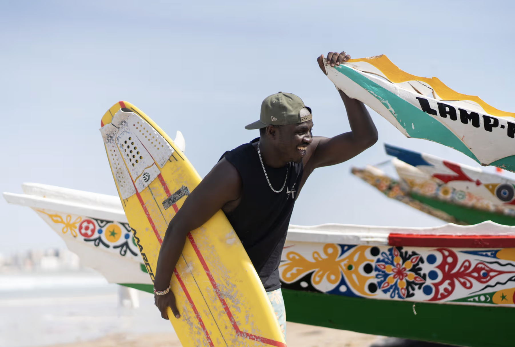 A man at the beach holding a yellow surfboard, smiling, with colorful decorated boats in the background.