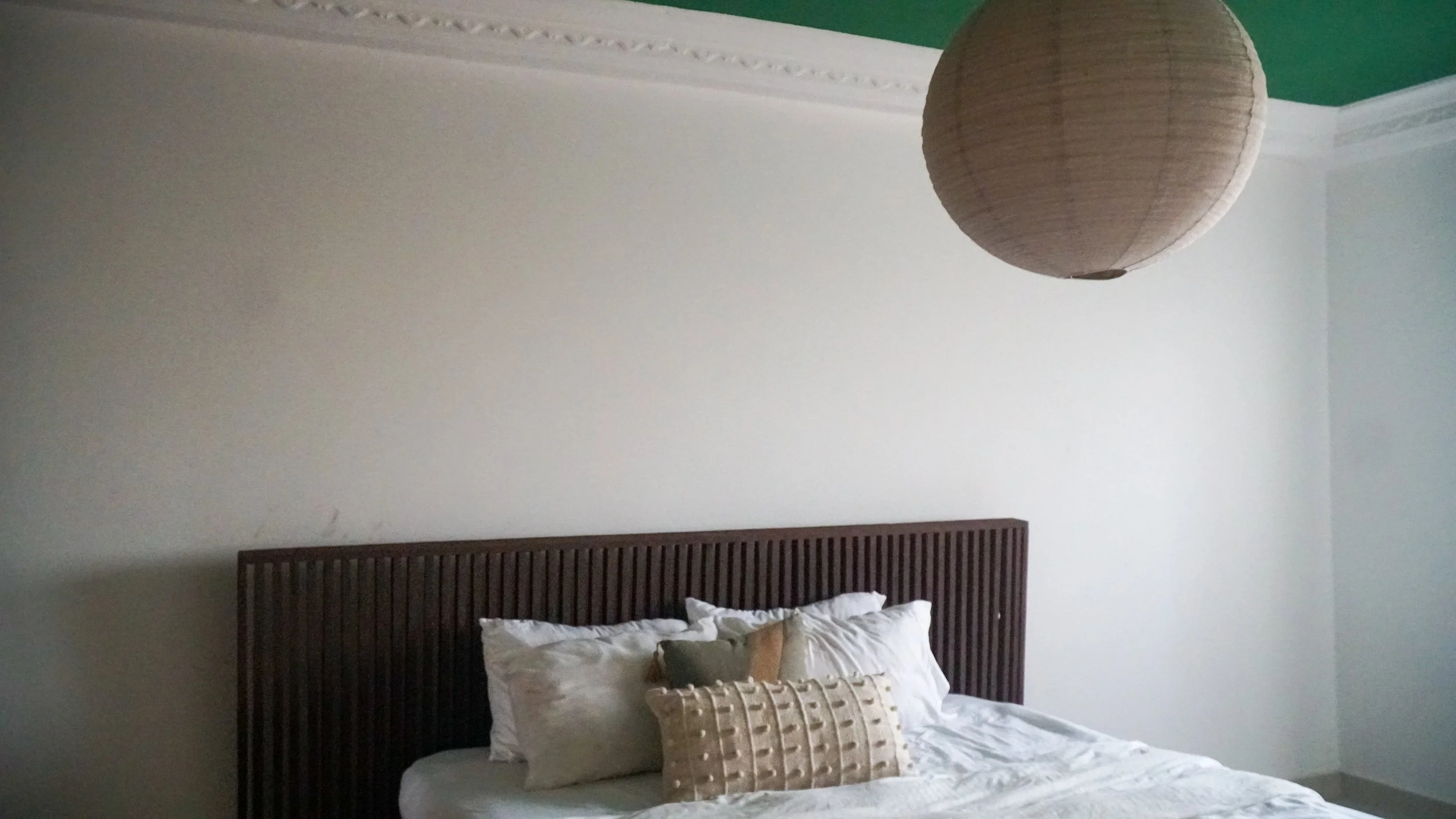 A bedroom with a white bed, dark wooden headboard, and decorative pillows. There is a large round paper lantern light fixture hanging from the ceiling. The walls are painted white with a green ceiling and decorative crown molding.