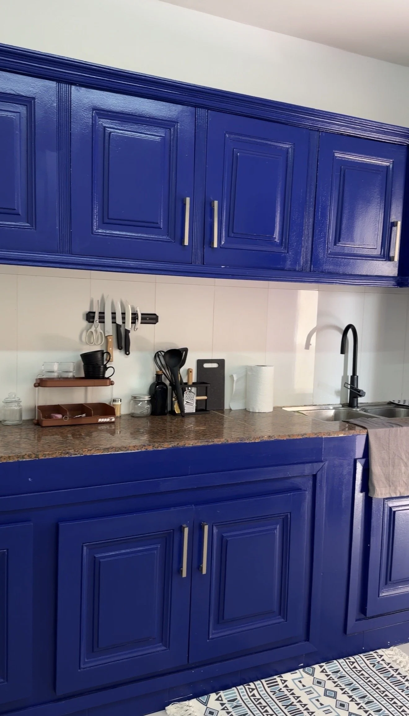 Kitchen cabinets painted blue, countertop with kitchen utensils, paper towel roll, sink and faucet, rugs on the floor.