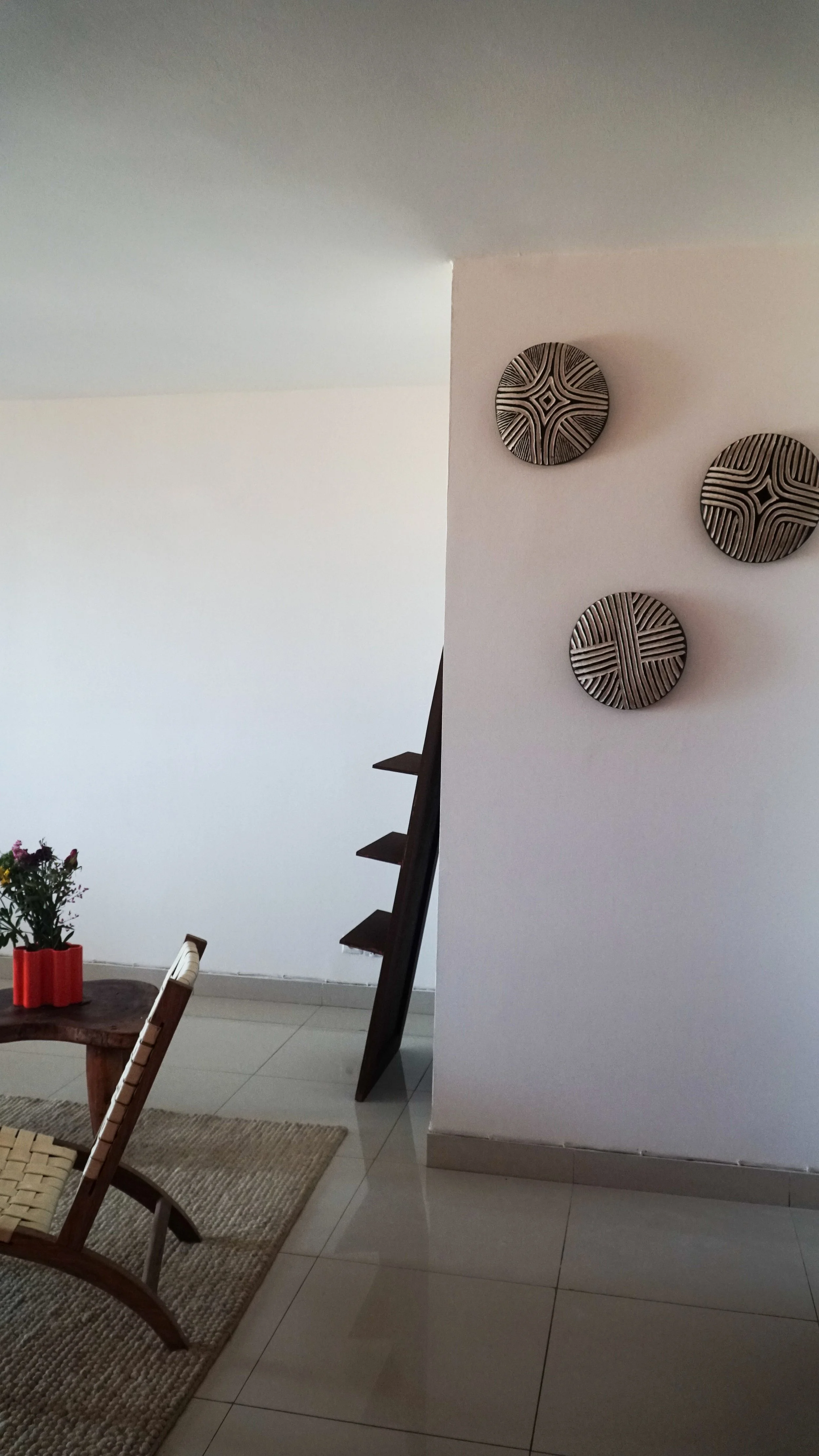 Interior of a house showing wall decor with three round patterned plates on a white wall, a wooden dining table with a flower vase, a woven chair, and part of a staircase.