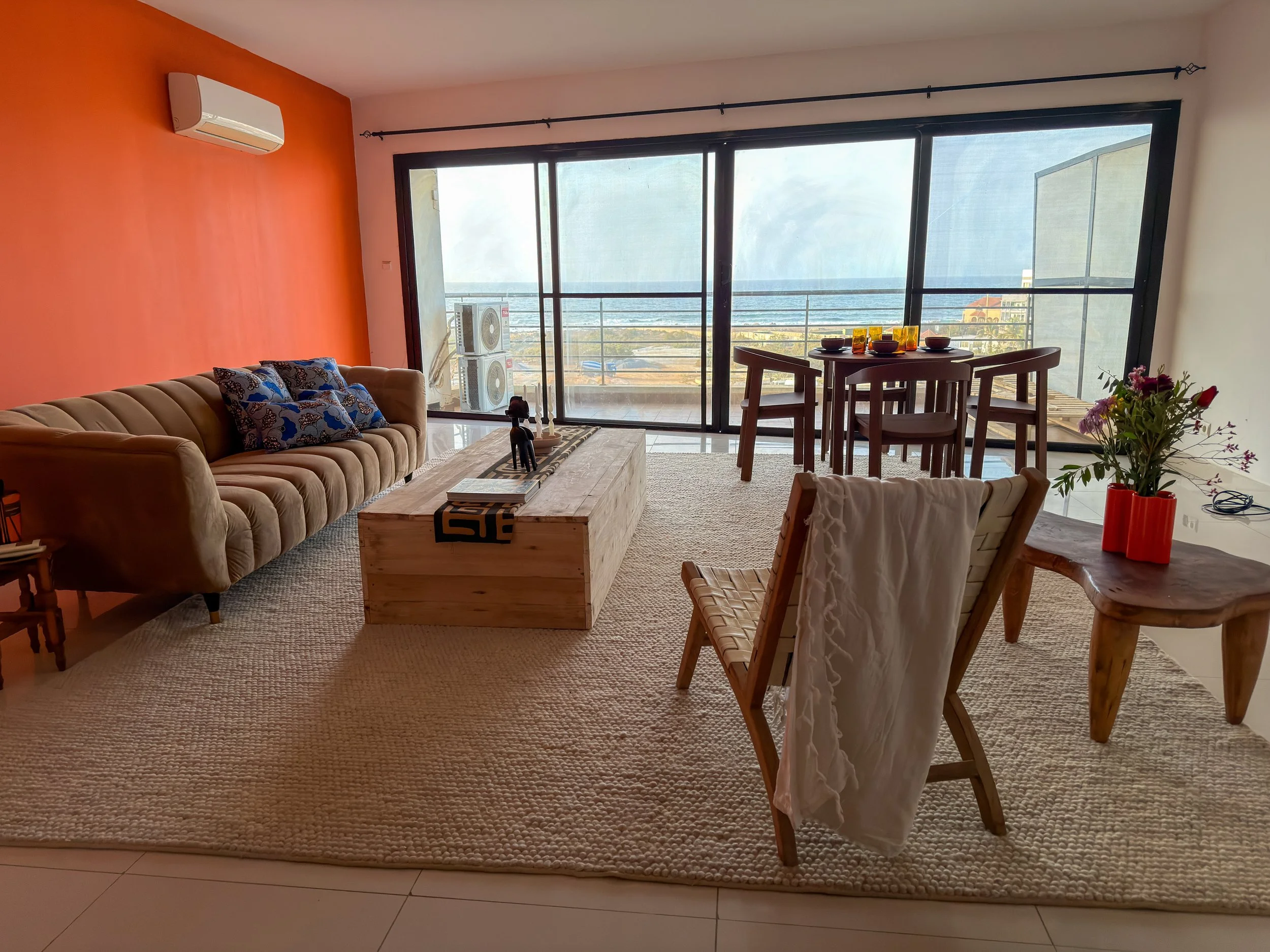 Living room with beige sofa, wooden coffee table, white armchair with a white throw, and a wooden dining table with four chairs near large balcony doors showing an ocean view. Decor includes colorful throw pillows, a vase with flowers, and wall-mount