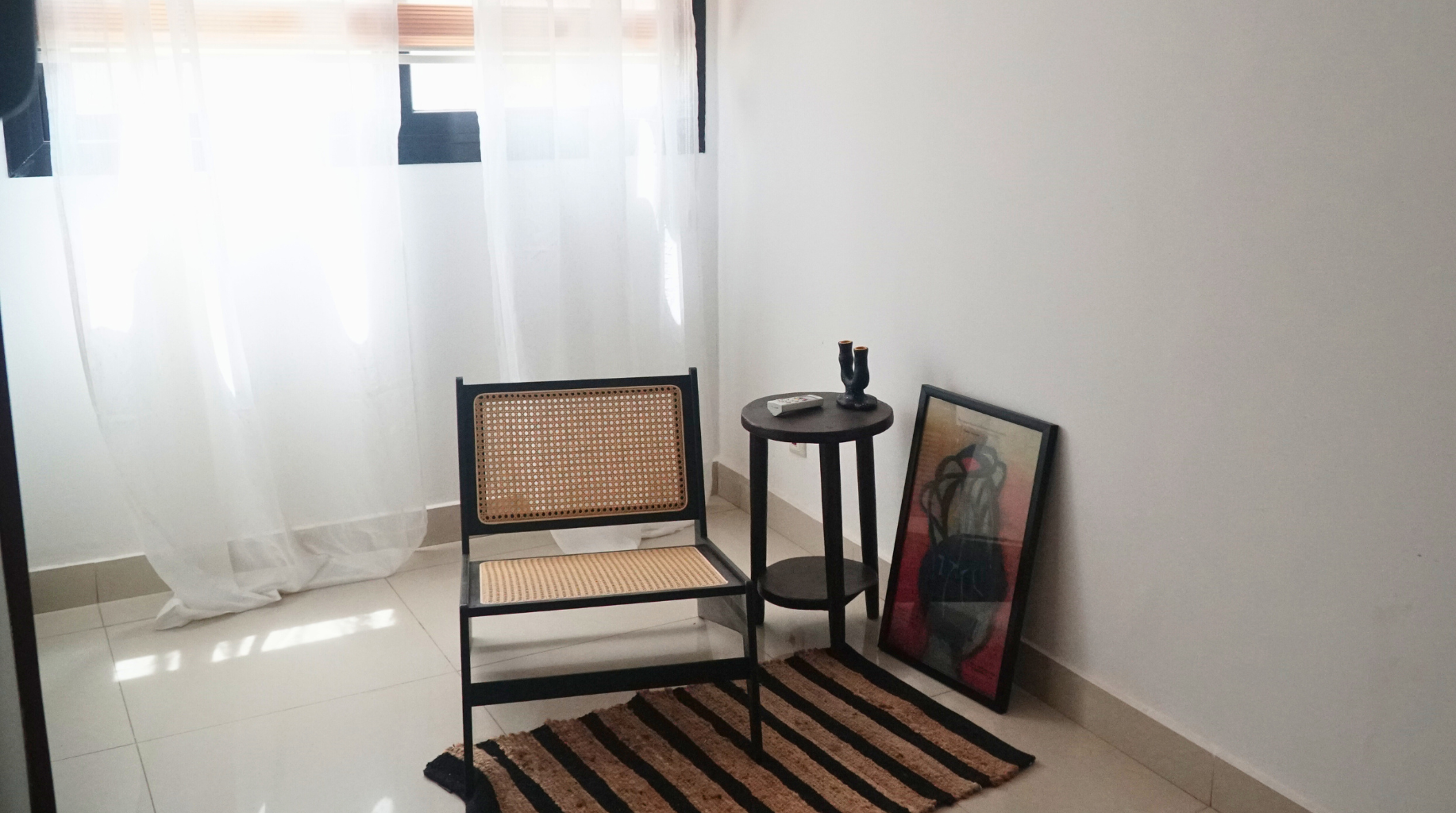 A small sitting area with a black chair with woven cane backrest and seat, a round black side table, a framed artwork leaning against the wall, a striped brown and black rug, and white sheer curtains covering a window.