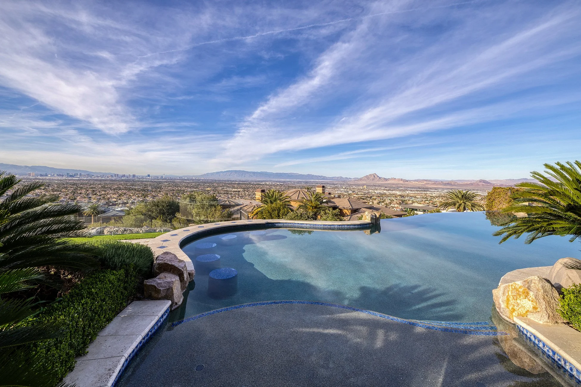 An infinity pool overlooking a residential area and distant cityscape with mountains in the background under a blue sky with wispy clouds.