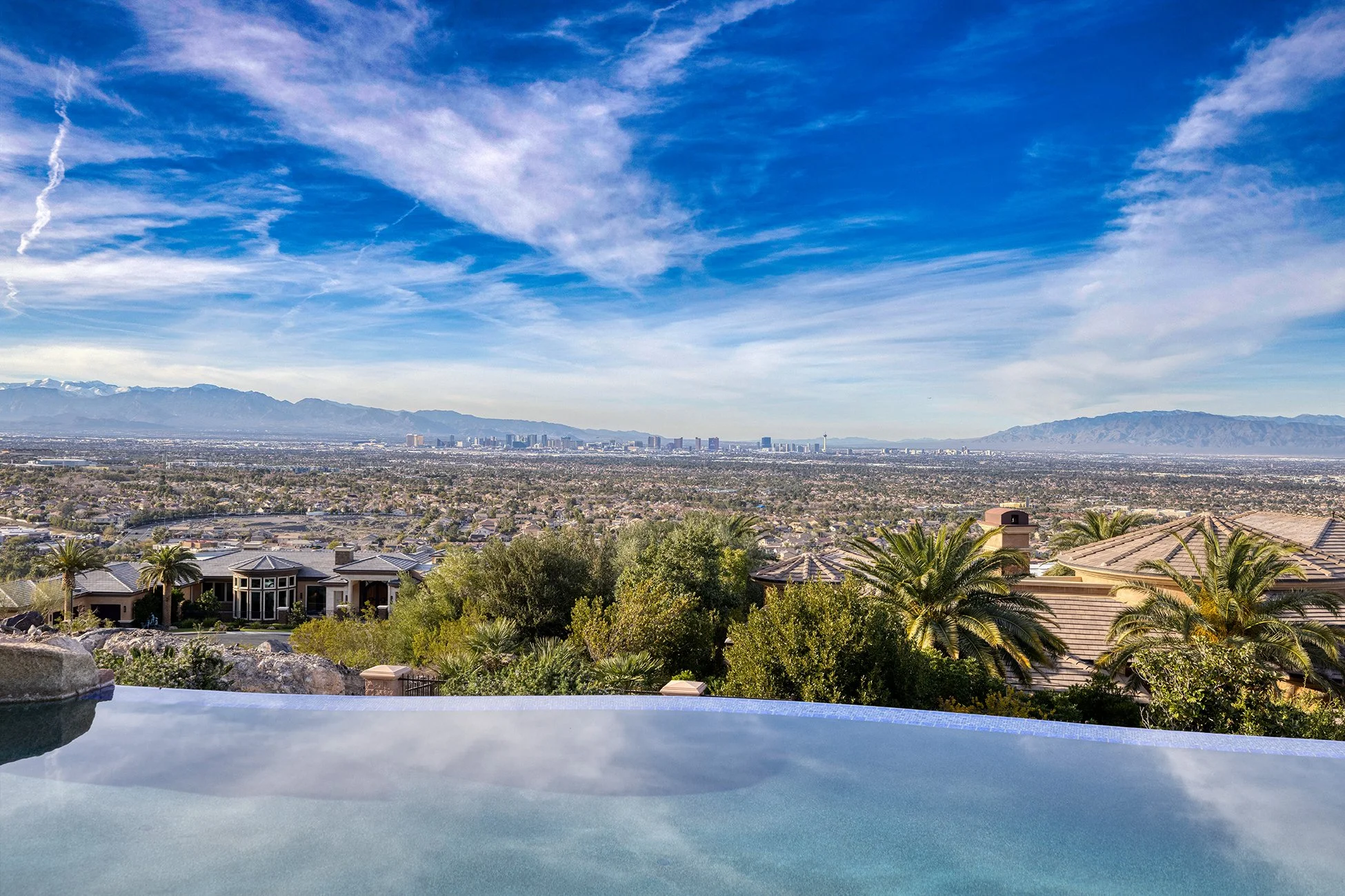 A scenic view from an infinity pool overlooking a residential neighborhood with desert landscaping, with mountains and a city skyline in the distance, under a partly cloudy blue sky.