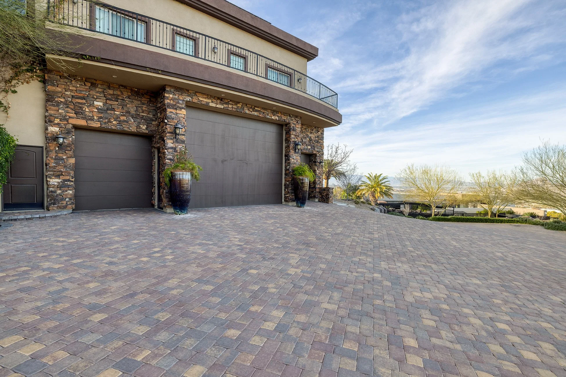 View of a modern house with a stone and stucco exterior, two garage doors, decorative planters, and a paved driveway in a landscaped area with trees and a clear sky.