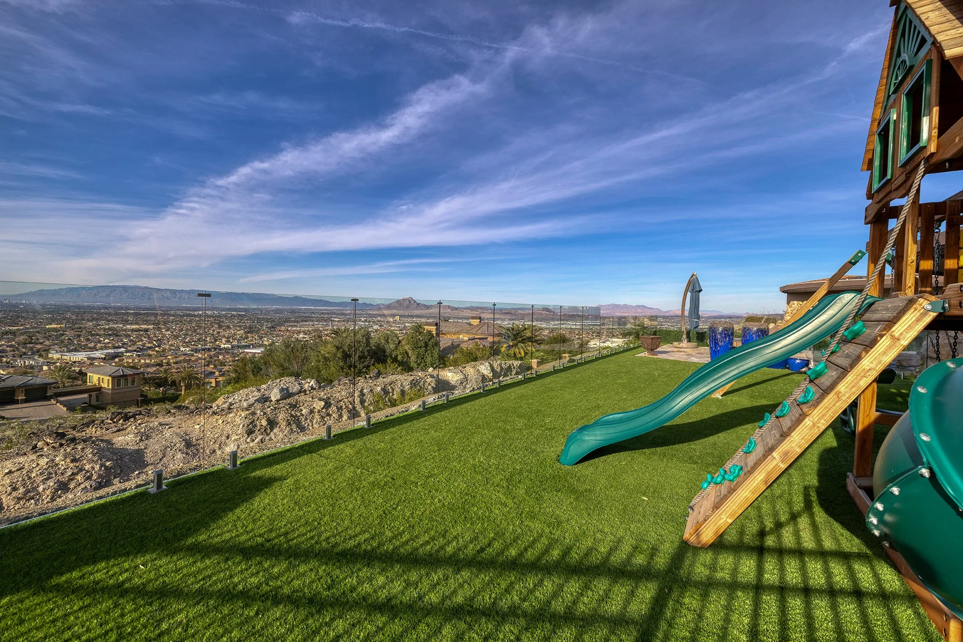 Backyard with artificial grass, playhouse with slide, rock landscape, city view, blue sky with clouds.