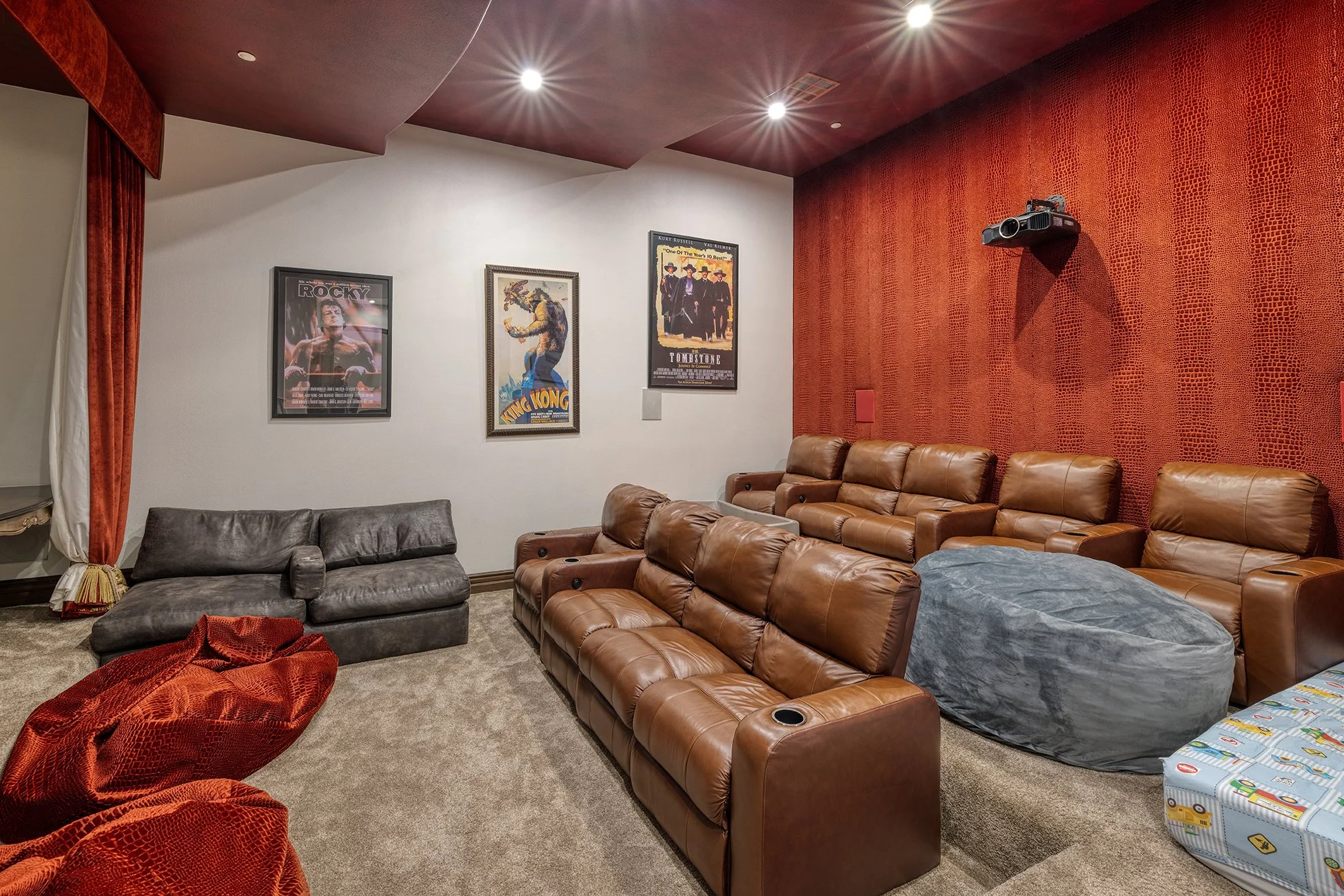 Home theater room with brown leather recliners, a gray bean bag, movie posters on the wall, a projector mounted on a red textured wall, and curtains on the side.
