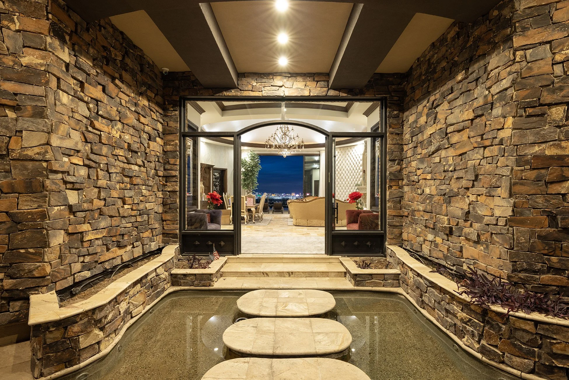 Interior view of a luxurious home with a stone accent wall, featuring a water feature with stepping stones leading into a spacious living room with elegant furniture and a chandelier, overlooking city lights at night.