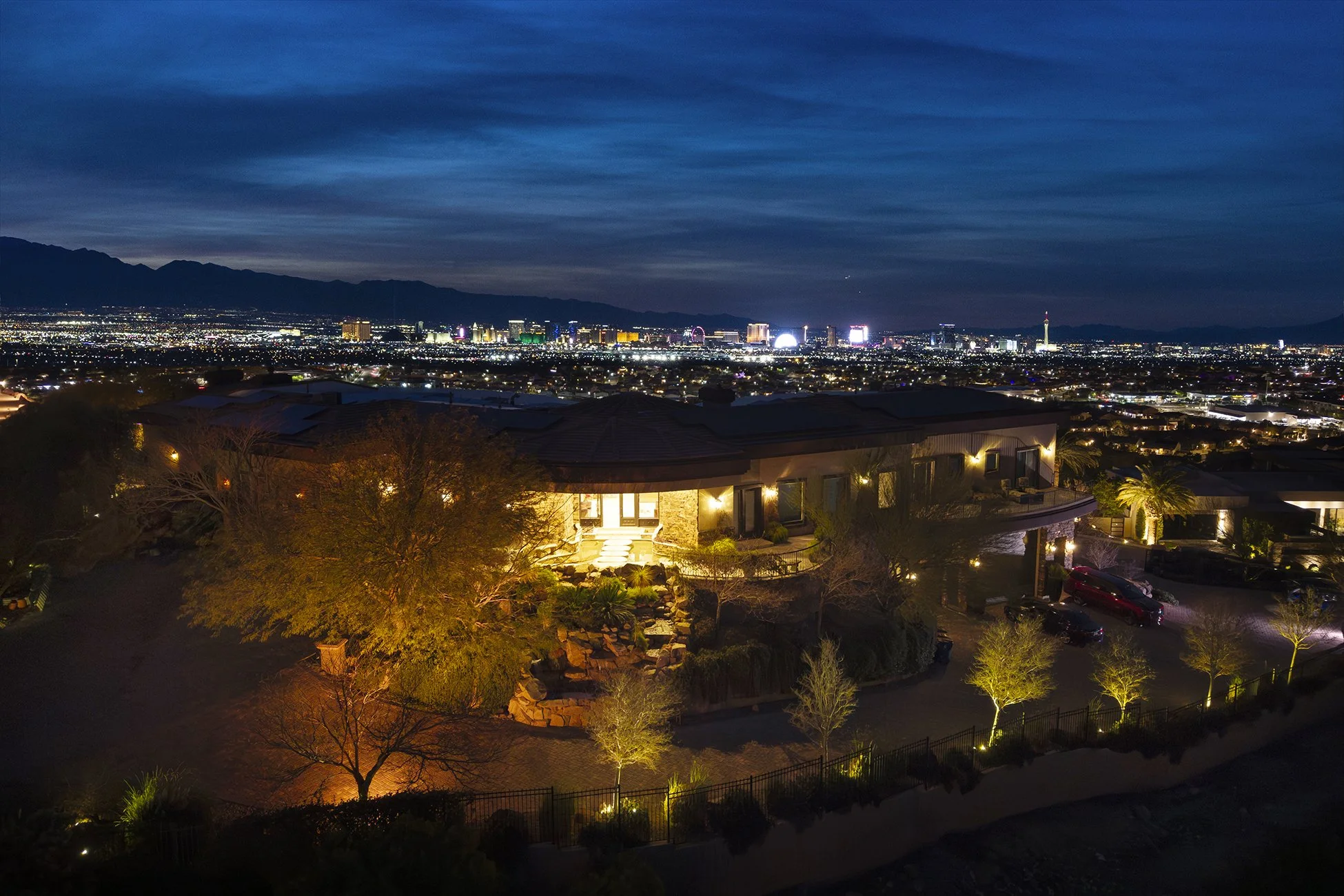 Nighttime view of a city skyline with illuminated buildings and mountains in the background, taken from a residential area with houses and trees lit by outdoor lighting.