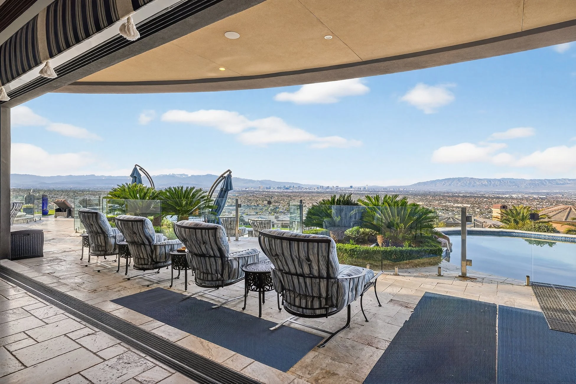 Luxury outdoor patio with striped cushioned chairs facing a swimming pool, overlooking a city skyline and mountains in the distance on a sunny day.