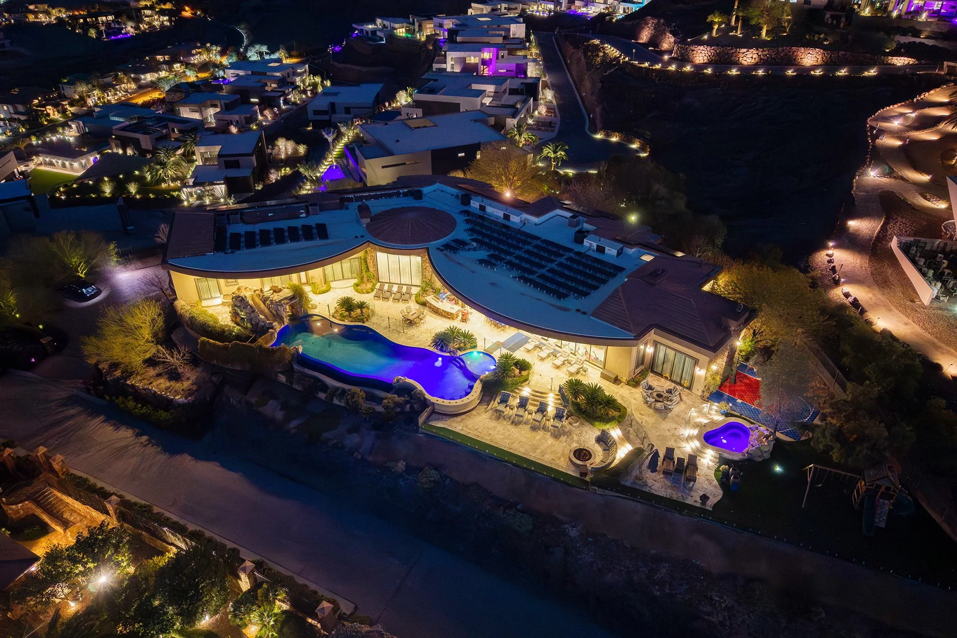 Aerial night view of a luxurious hillside mansion with illuminated pools, outdoor seating, and tropical landscaping, surrounded by other brightly lit upscale homes.