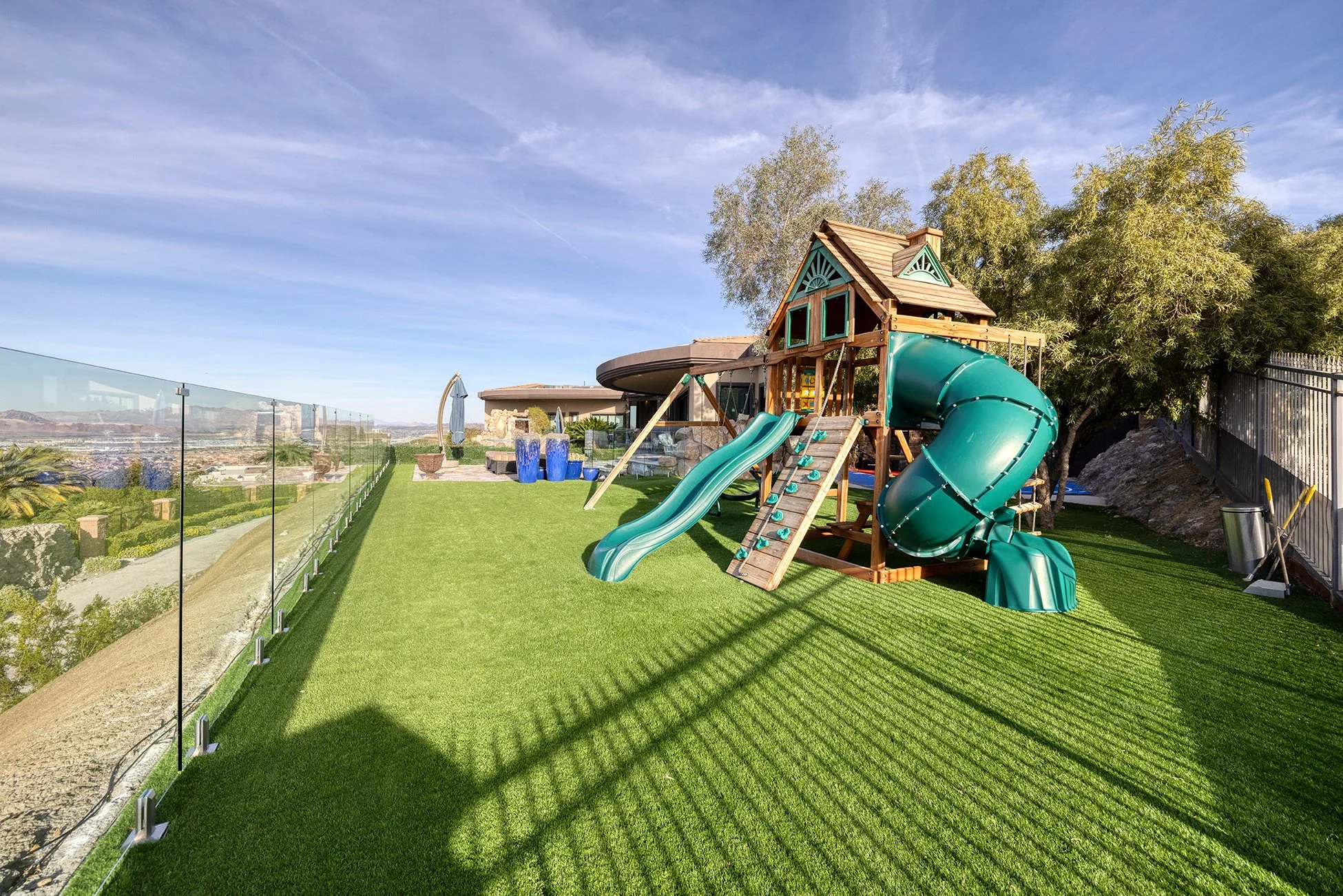 Backyard with artificial grass, a wooden playset with slides, climbing wall, and tower, a glass fence, trees, and a blue sky.