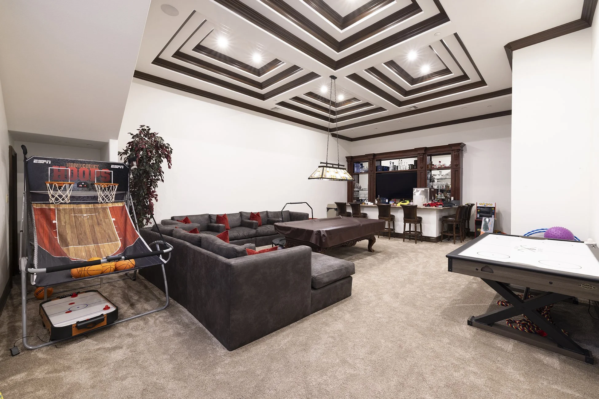 A spacious basement entertainment room with white walls, a decorative ceiling with recessed lighting, and carpeting. It features a large sectional sofa with red pillows, a billiards table, an air hockey table, and a bar area with wooden stools. There