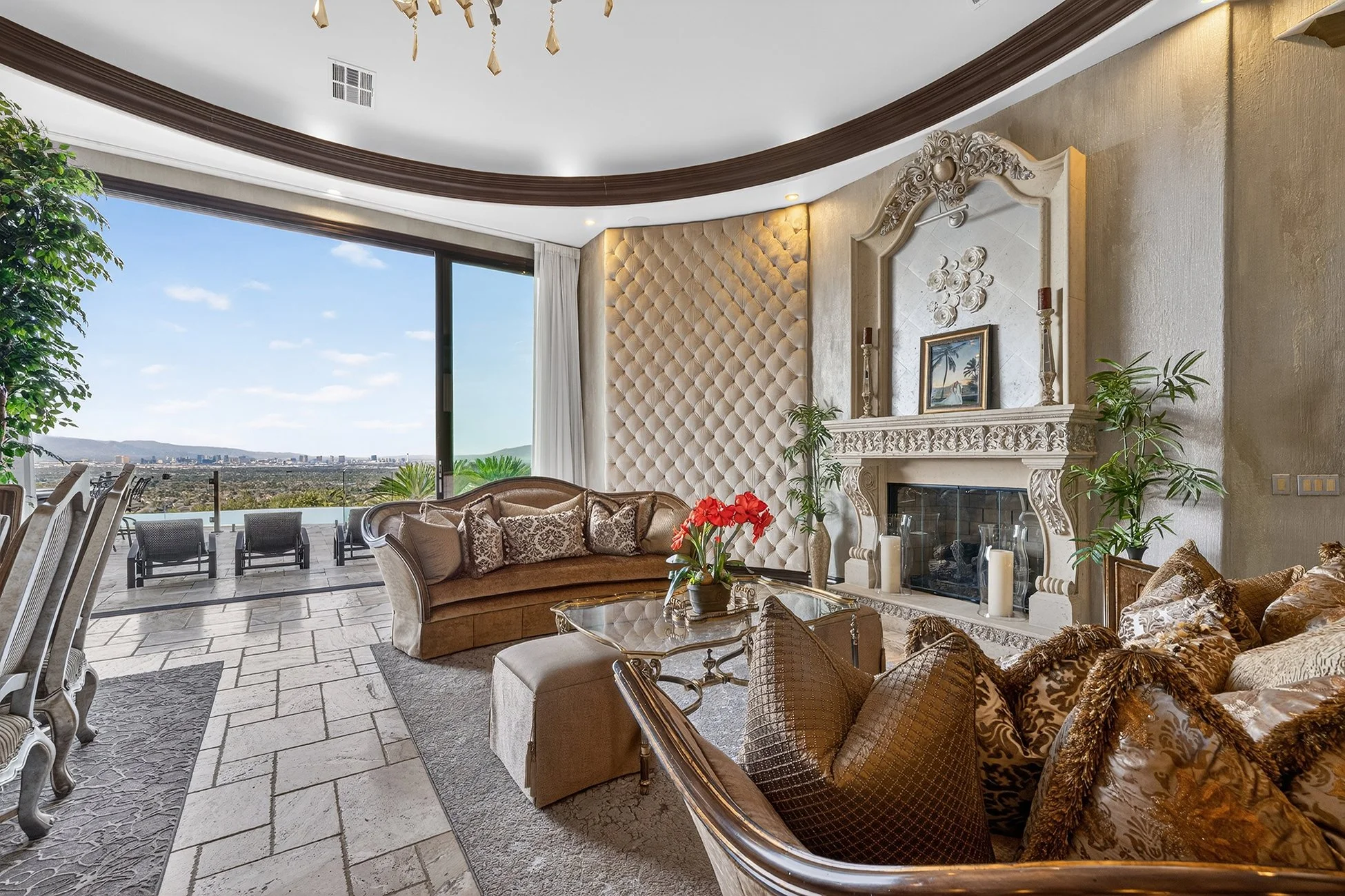 Elegant living room with large balcony and city view, tufted wall, ornate fireplace, plush sofas, colorful pillows, glass coffee table, and decorative plants.
